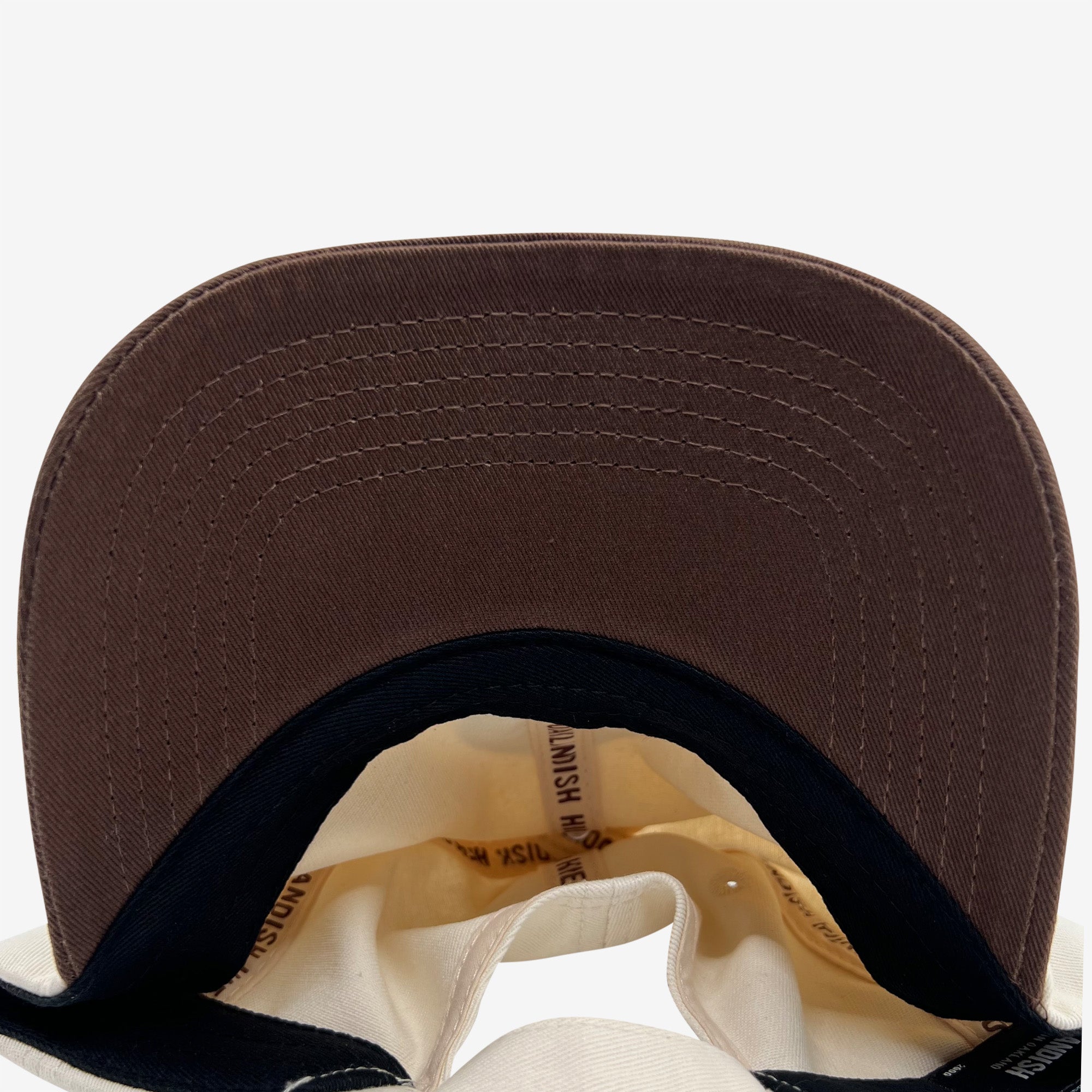 A close-up view of the underside of a Hieroglyphics Hiero Logo Dad Hat's brim, highlighting the detailed stitching. The inside label features printed text on a beige fabric. The interior of the cap is lined with black and beige fabric, showcasing the underside view and construction details.