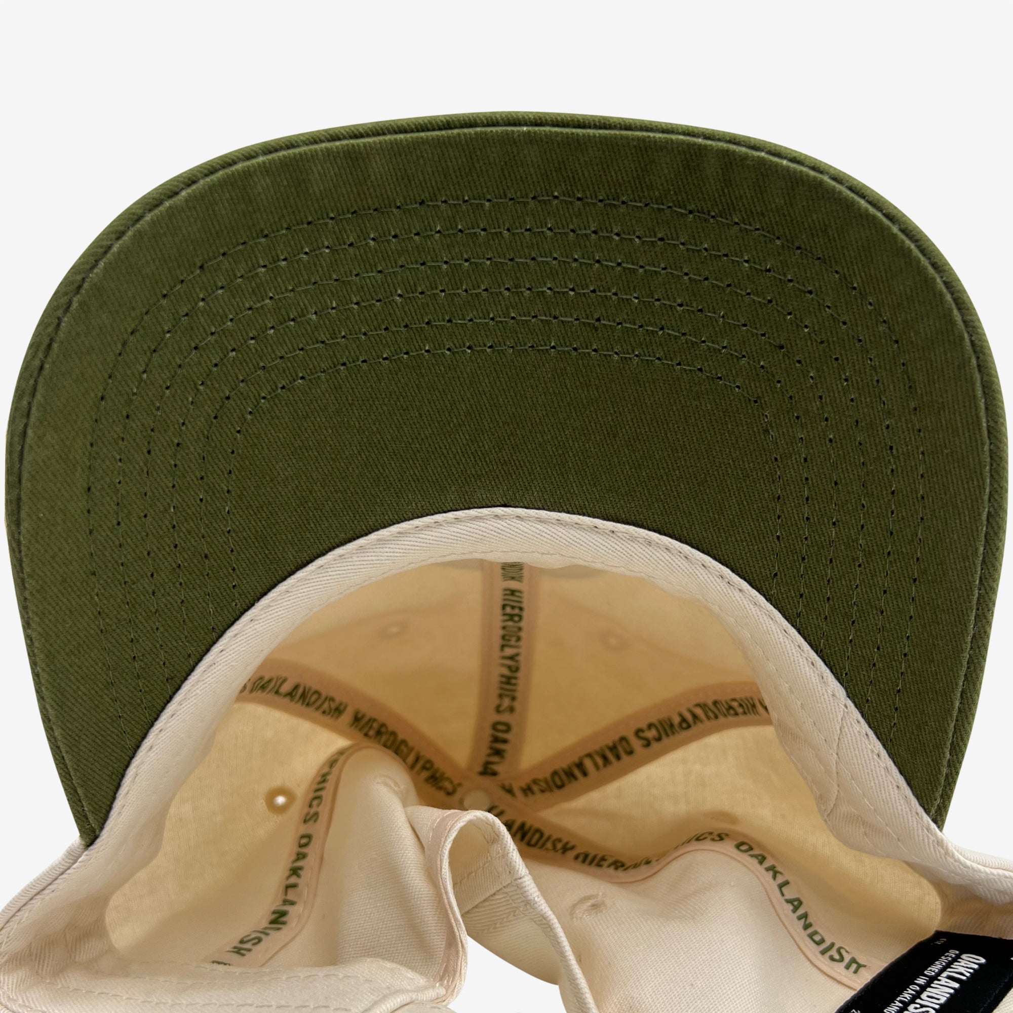 The image displays the underside of a green Hiero Logo Dad Hat from Hieroglyphics with a beige inner lining. The hat's brim shows detailed stitching in matching green thread. Inside, there is text along the seams that reads "OAKLANDISH MERCH" repeatedly.