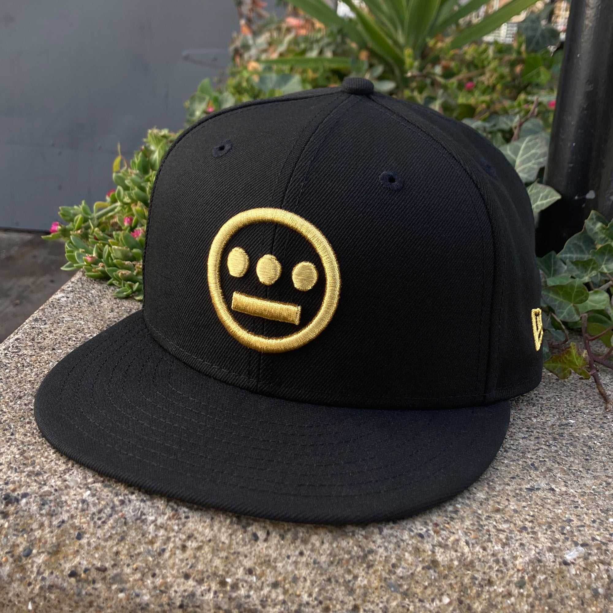 The New Era Hiero 59FIFTY Fitted Cap by Hieroglyphics is a black cap with a flat brim, adorned with a distinctive yellow embroidered logo featuring three dots for eyes and a horizontal line for a mouth, all enclosed within a circle. Positioned on a concrete surface against the backdrop of green foliage and pink flowers, the cap evokes the essence of modern hieroglyphics.