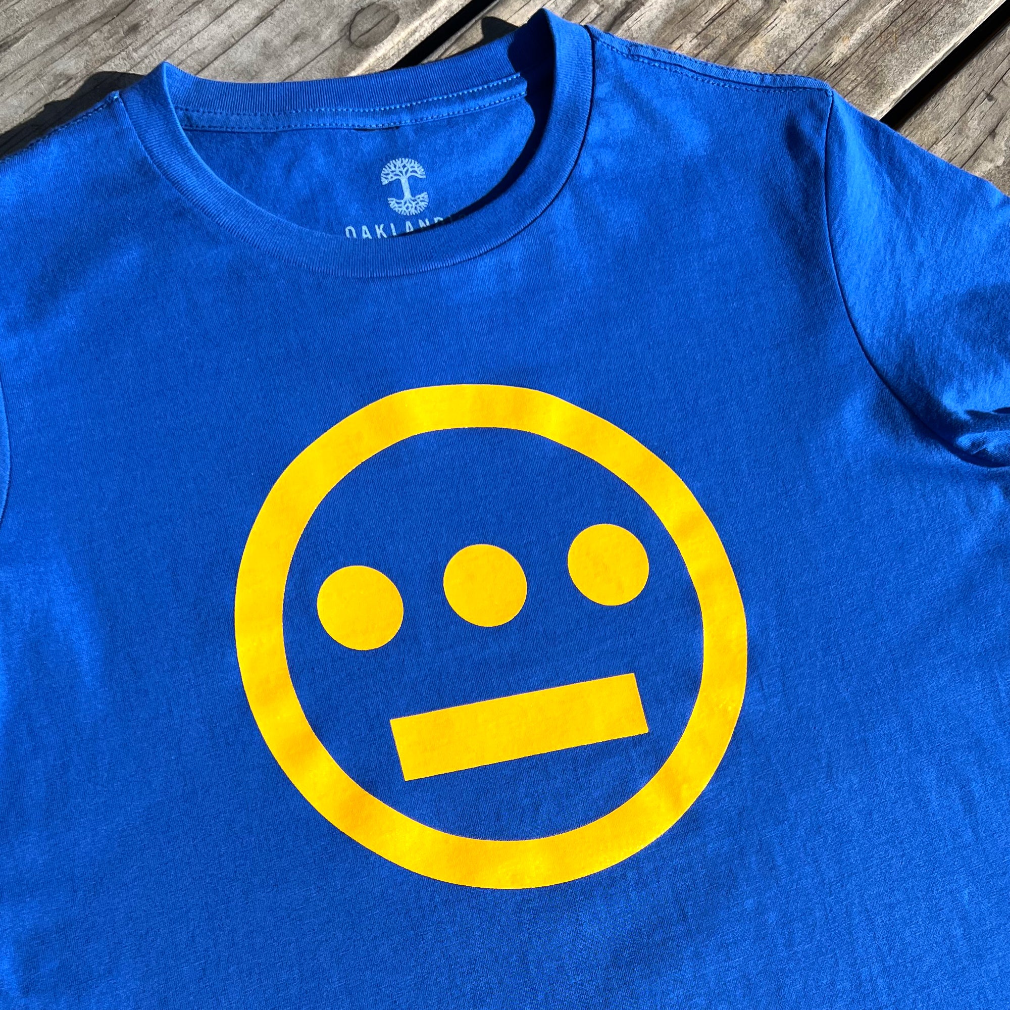 This image features the Women's Hiero Classic Logo Tee by Hieroglyphics. The royal blue t-shirt showcases a yellow logo with a circle enclosing three equally spaced dots in a horizontal line above a separate horizontal line at the bottom, reminiscent of hieroglyphics. The t-shirt is displayed on a wooden surface, and part of the neckline label is visible.