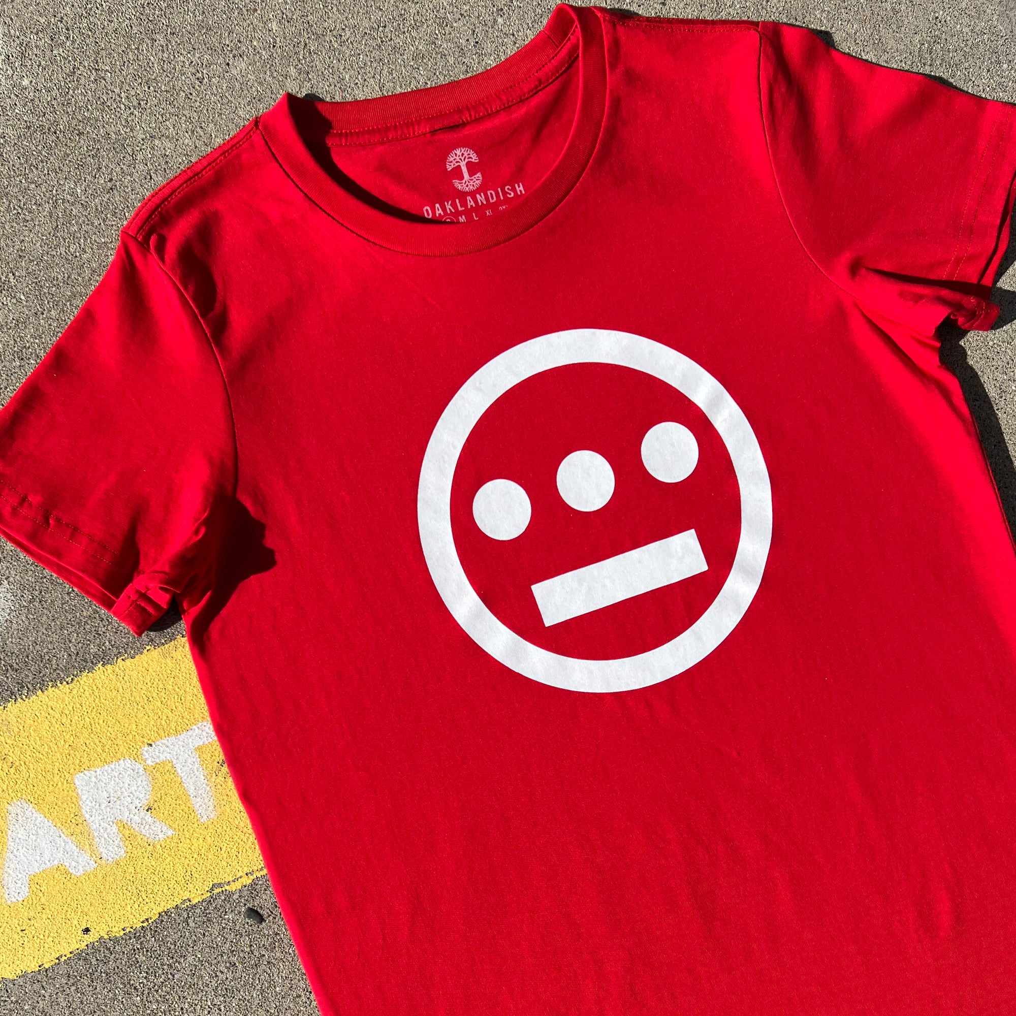 The Women's Hiero Classic Logo Tee by Hieroglyphics, a red 100% cotton T-shirt, is displayed laid flat on a concrete surface. It features a white minimalist design on the front, consisting of a circle with three dots forming a horizontal line near the top and a single horizontal line near the bottom, reminiscent of hieroglyphics. The T-shirt tag reads "OAKLANDISH.