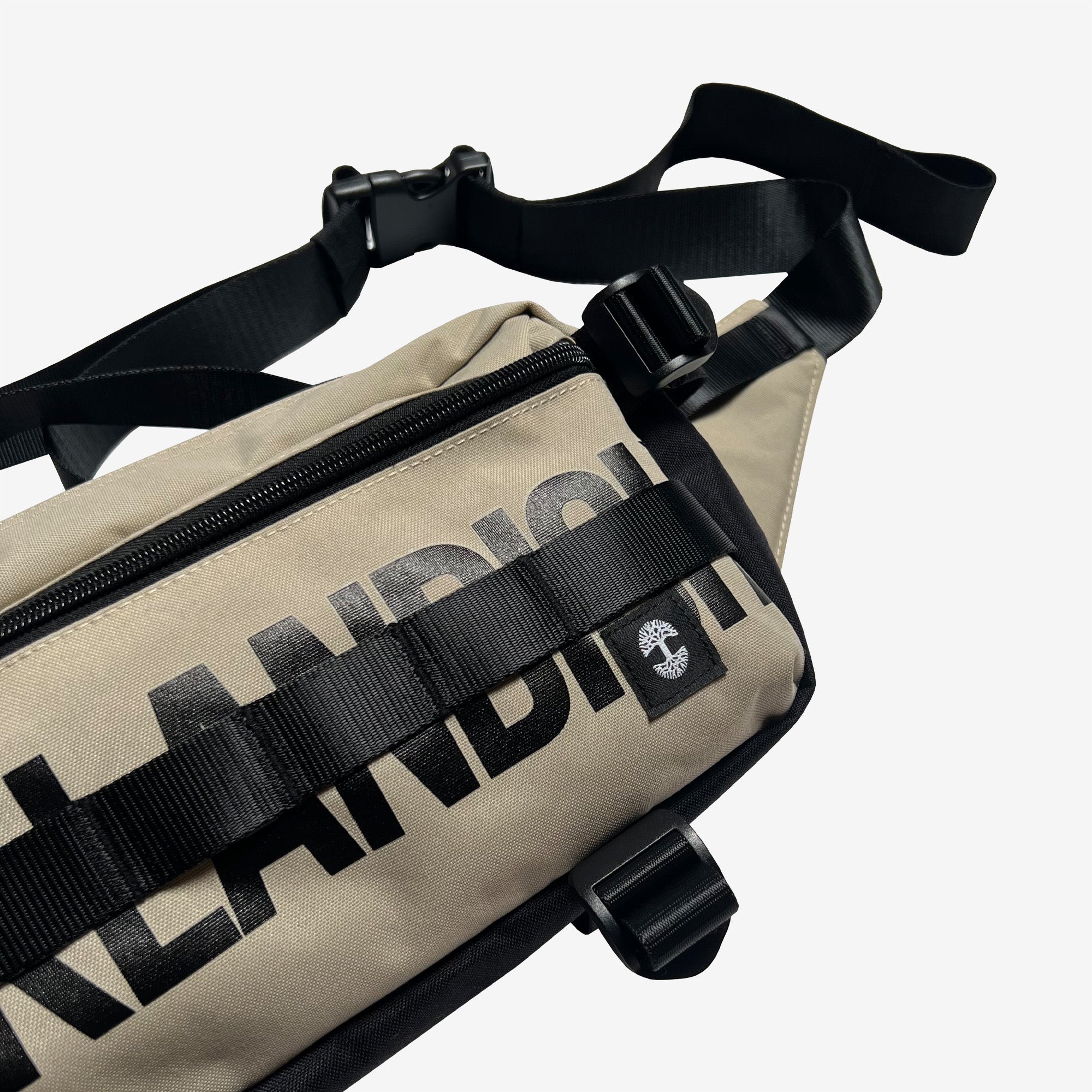 Close-up of the Oaklandish Hip Bag in beige with black zippers and straps. The front features bold, black letters with partial text visible, ending with "NDIS," along with a small tree logo. This versatile hip/shoulder bag is set against a plain white background.