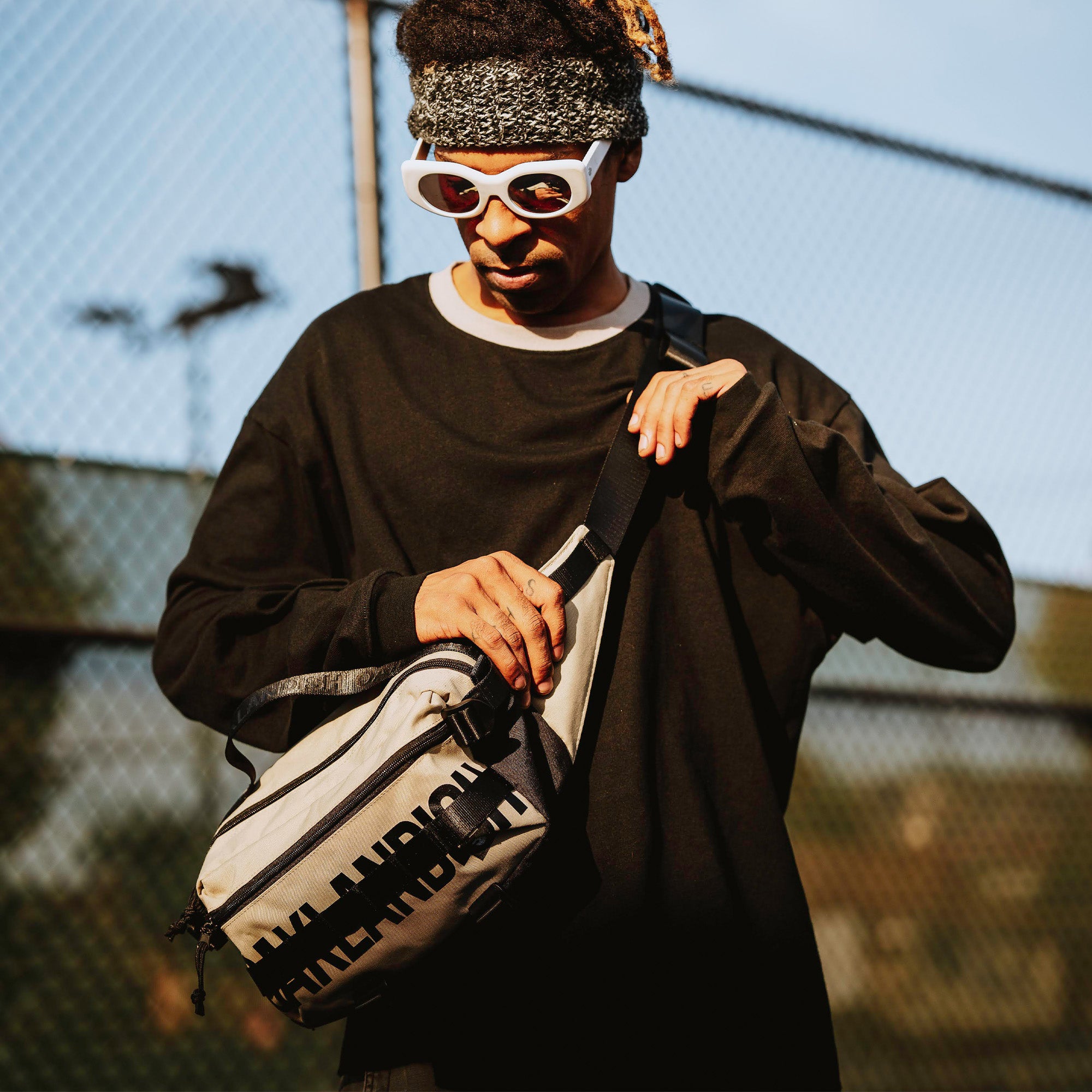 A person stands outdoors against a blurred chain-link fence background, wearing a loose black long-sleeve shirt and white, futuristic sunglasses. They sport a grey knit headband and carry an Oaklandish Hip Bag with black text. The individual adjusts the strap of their Oaklandish bag with one hand.