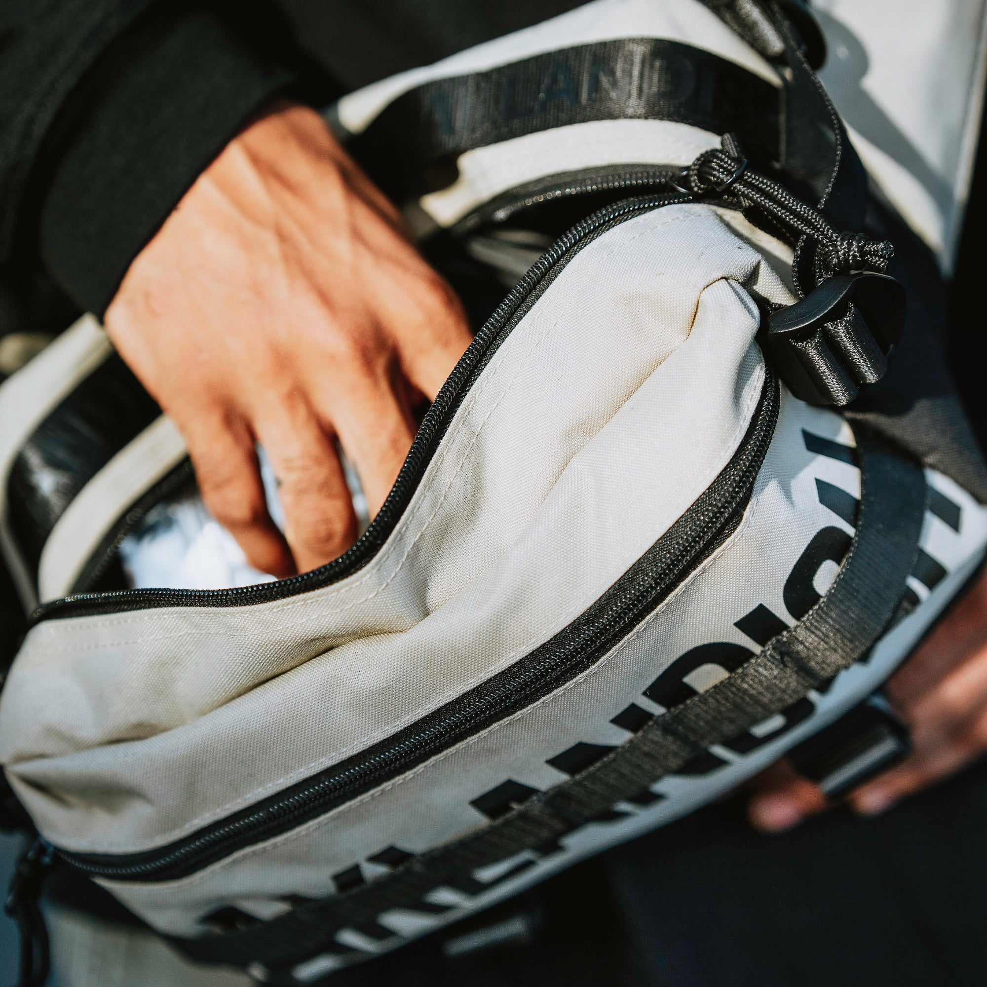 Close-up view of a person opening an Oaklandish Hip Bag with the strap slung over their shoulder. The person's hand is reaching into the main compartment of the bag, which has a partially open zipper. The Oaklandish bag appears worn, with prominent black text partially visible on its surface.