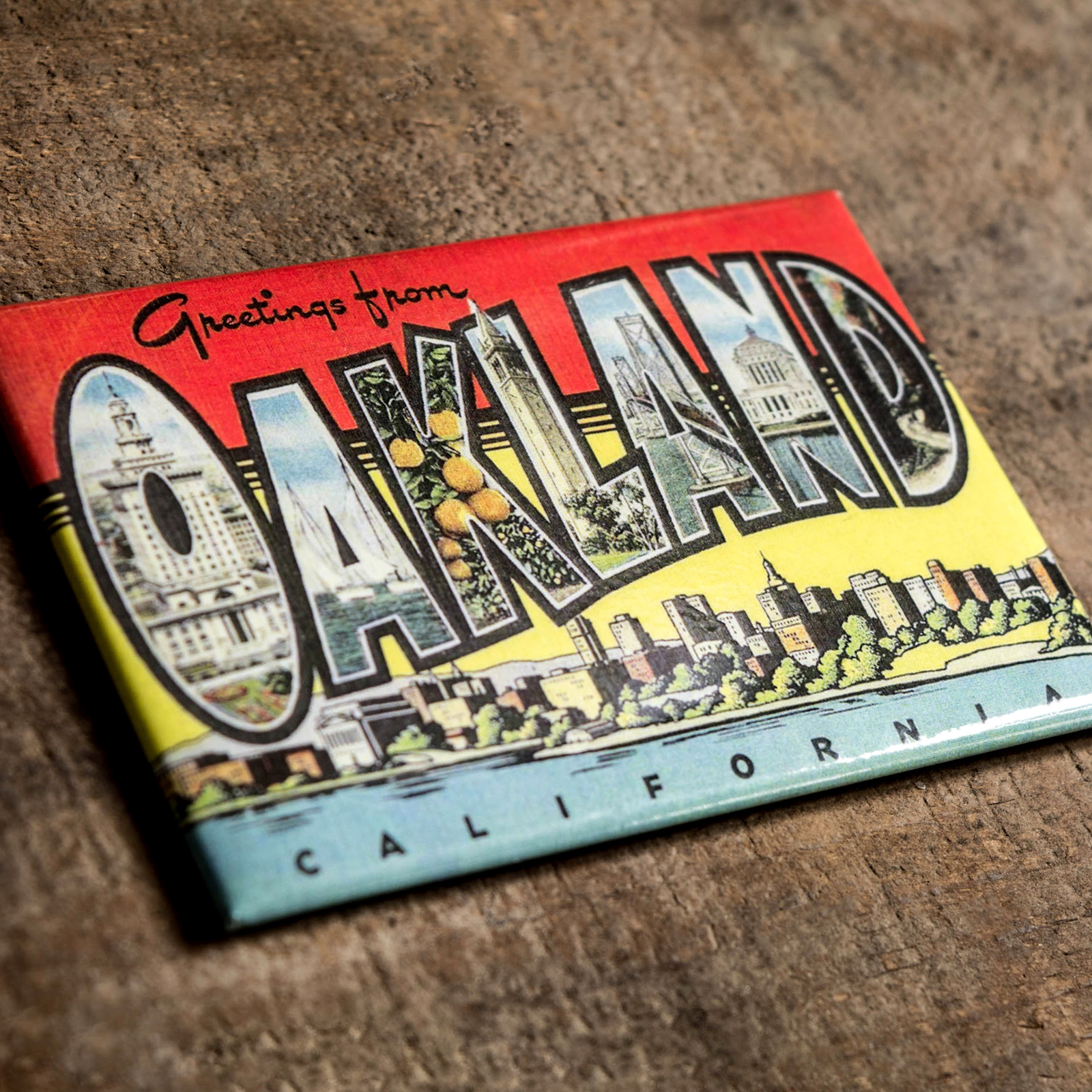 A collectible refrigerator magnet from Oaklandish lies on a textured surface. The item, named "Greetings from Oakland Magnet," features the words "Greetings from OAKLAND CALIFORNIA" in large, bold letters. Each letter of "OAKLAND" contains images of city landmarks and scenery, against a colorful gradient background that transitions from red to yellow.