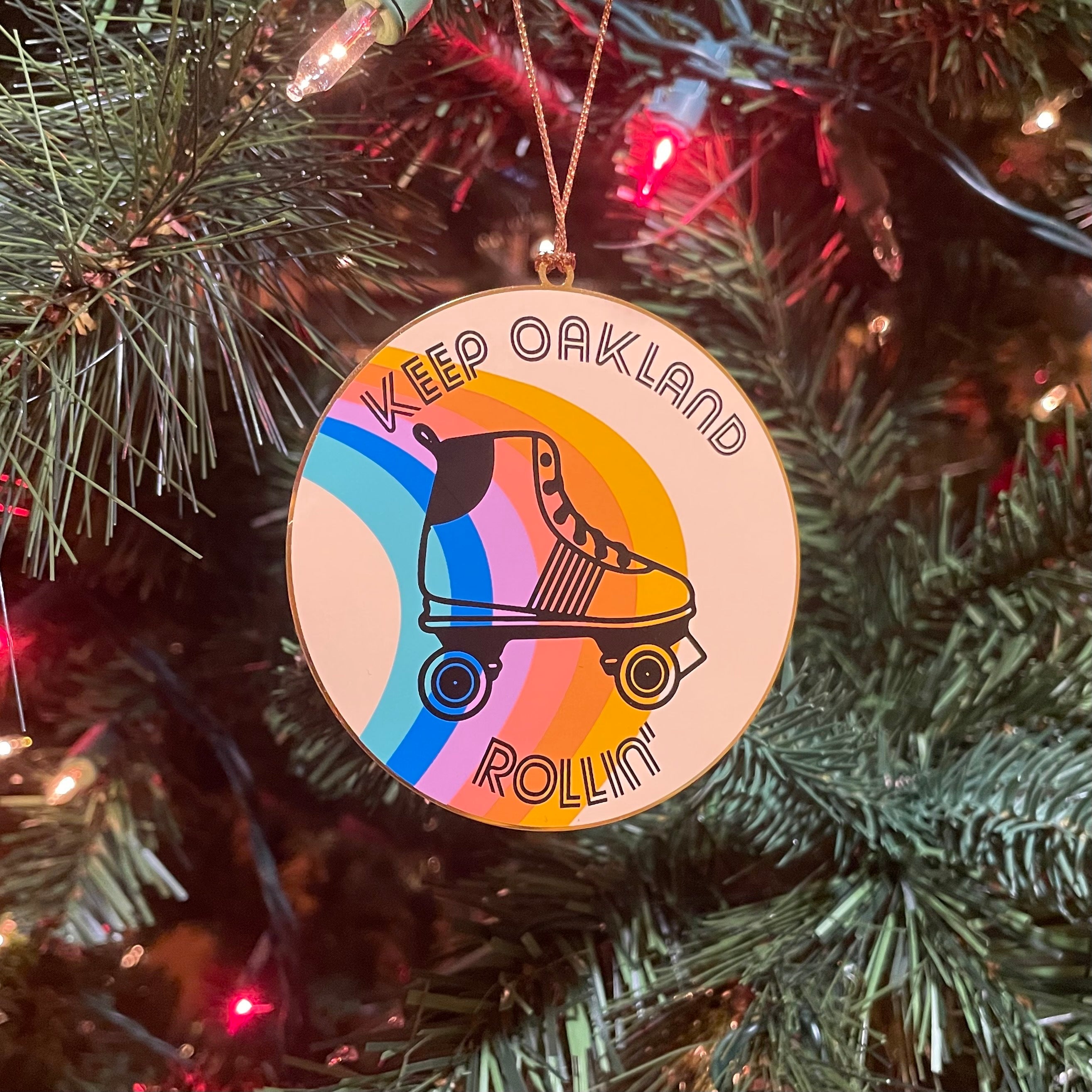 Keep Oakland Free holiday tree ornament with an illustration of a roller skate on a muted rainbow-colored circular disc hanging from a Christmas tree.