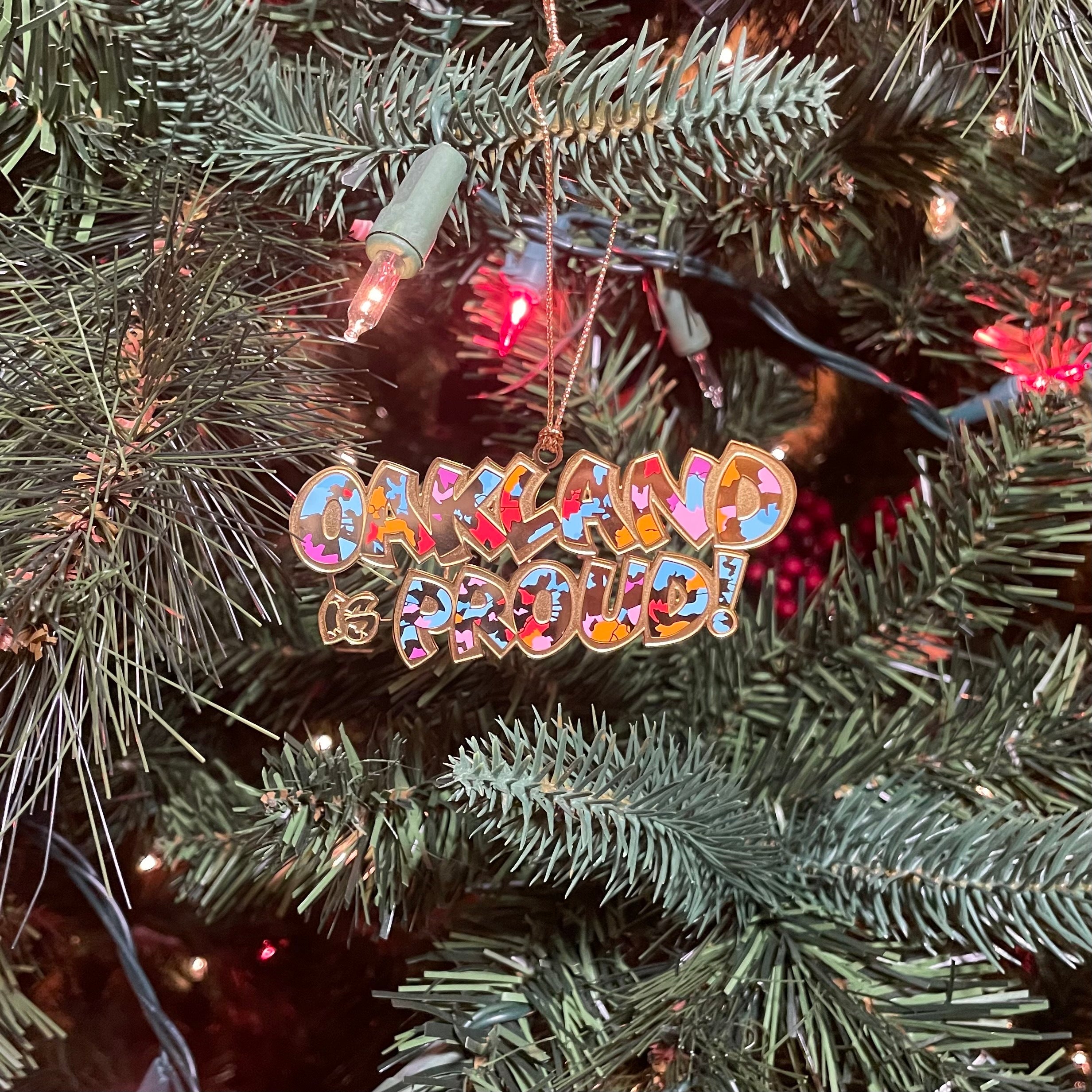 Oakland is Proud full-color wordmark depicted in cut-out brass holiday ornament hanging on a decorated Christmas tree.