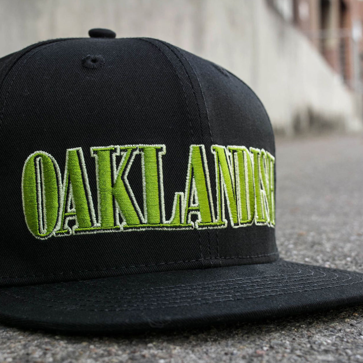 A close-up image of the Greenback Snapback by Oaklandish, featuring the word "OAKLANDISH" embroidered in bold, green letters on the front. The black snapback cap is placed on a rough concrete surface, slightly angled to the right corner. The background showcases a blurred urban setting with industrial buildings.