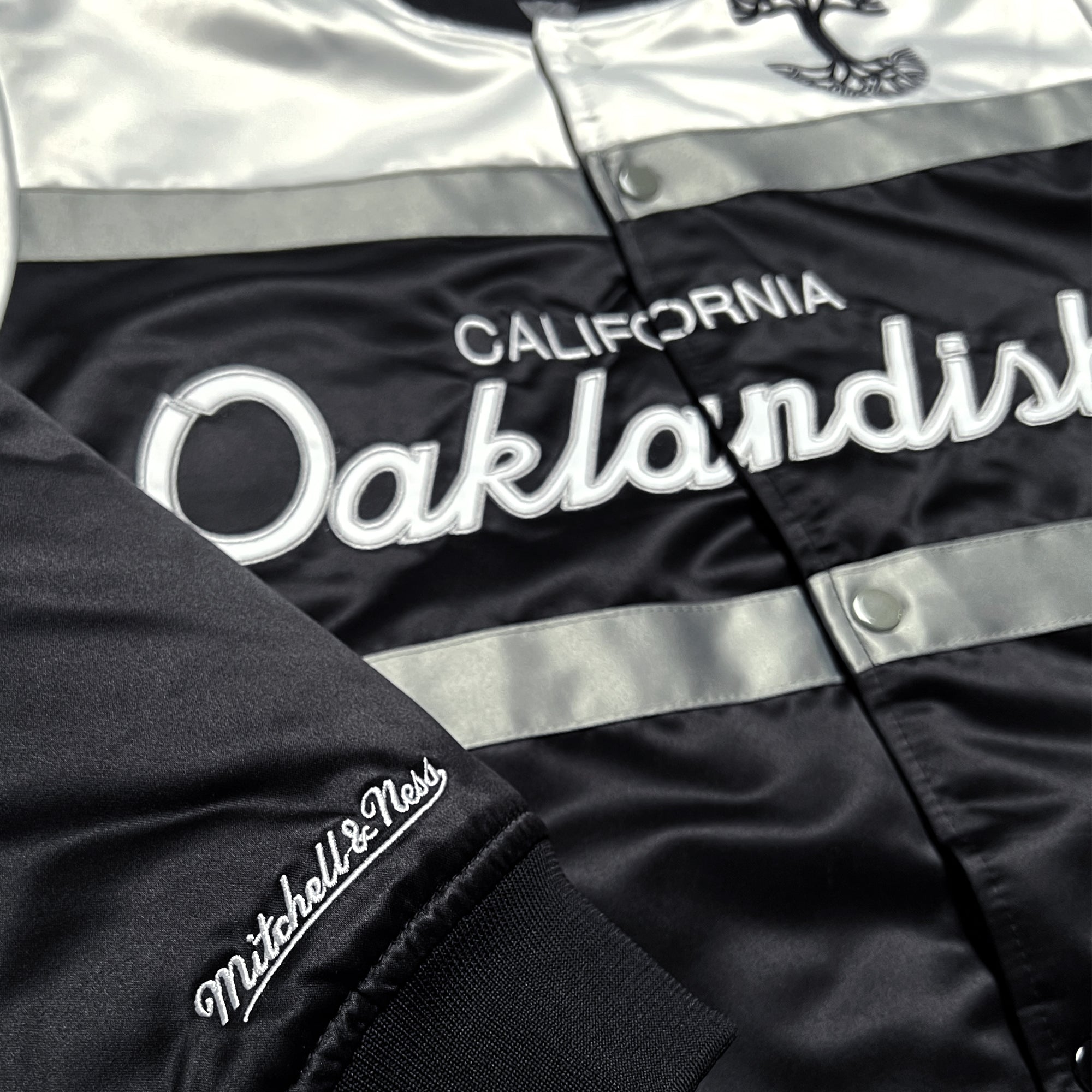 Close-up of Mitchell & Ness wordmark on the sleeve and California Oaklandish wordmark on the chest of a striped satin jacket.