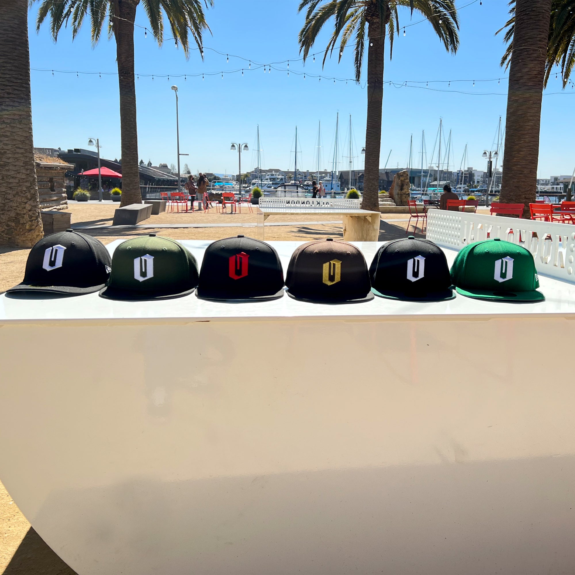 A row of six vibrant New Era Official 59FIFTY Fitted Caps is displayed on a shiny white surface outdoors. Each piece of headwear features a distinctive logo, including several from Oaklandish. Two palm trees, string lights, red chairs, and a marina with docked boats complete the scenic backdrop under a clear blue sky.