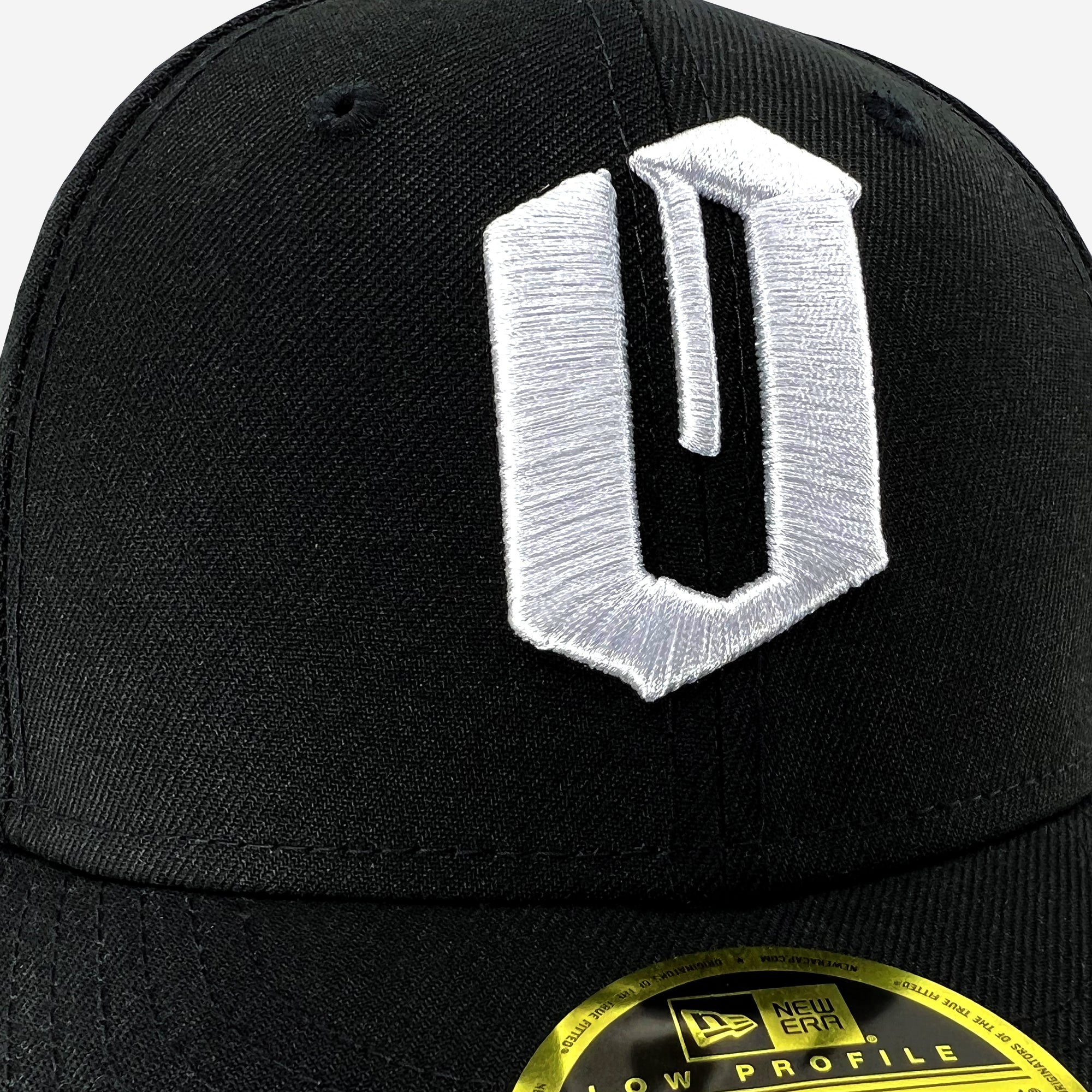 Close-up image of a black low-profile New Era Official 59FIFTY Fitted Cap by Oaklandish, featuring a large, white, embroidered letter "U" on the front. A circular gold and black sticker near the brim displays the 59FIFTY brand logo and information on the hat's style. The image highlights the hat's texture and stitching details.