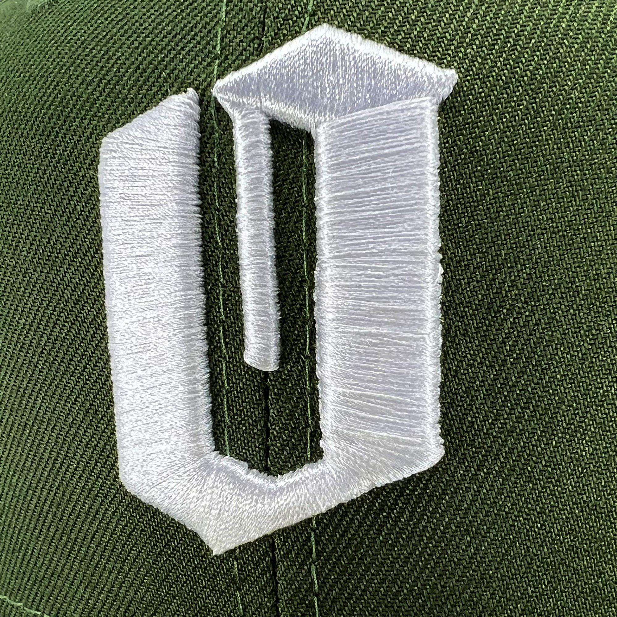 Close-up of a green fabric with a white embroidered design. The design features an abstract, angular shape resembling an incomplete letter "U" or "C," prominently displayed with clean stitching. The craftsmanship of the New Era Official 59FIFTY Fitted Cap by Oaklandish is emphasized by the detailed stitching and visible fabric texture.