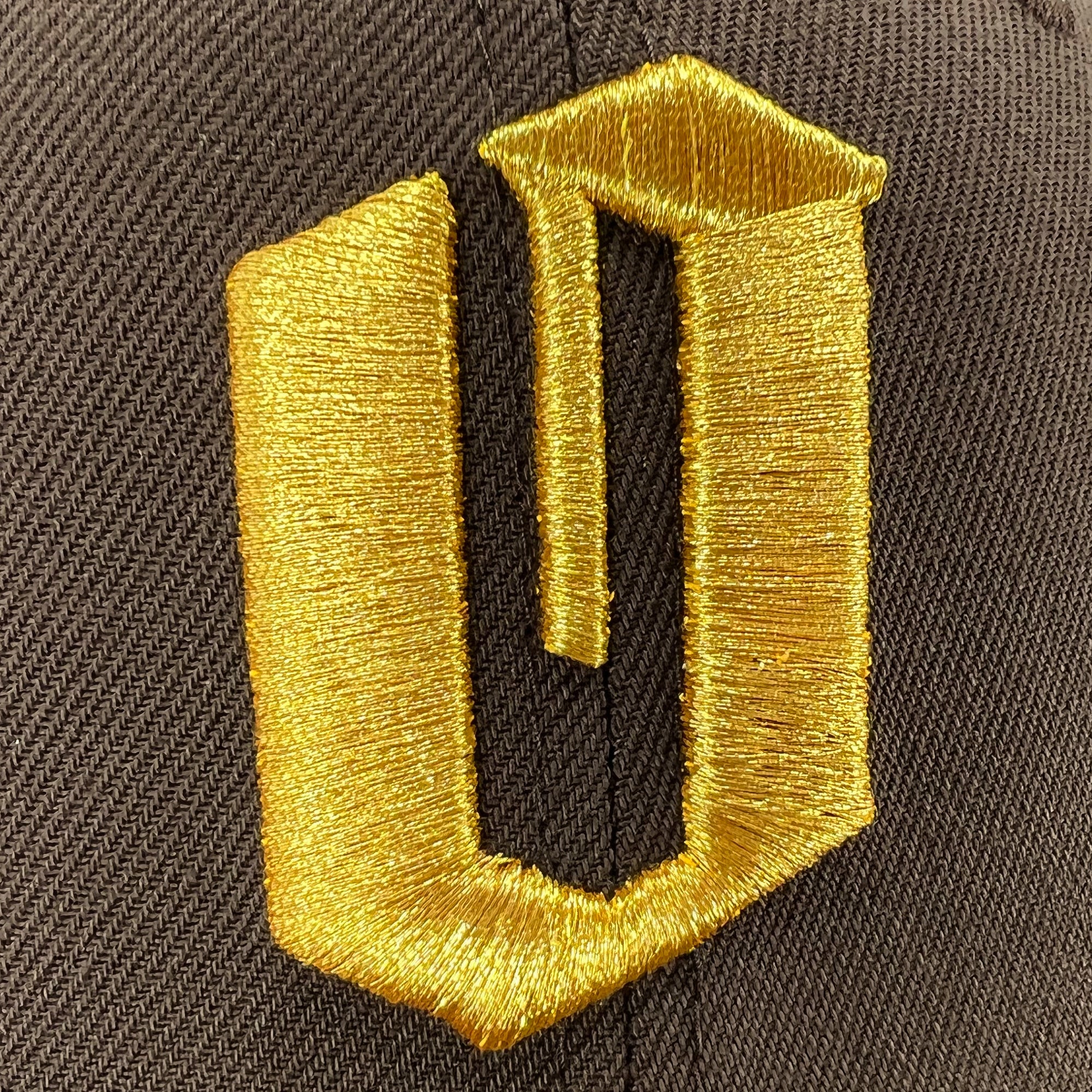 A close-up image of a black New Era Official 59FIFTY Fitted Cap featuring a prominent gold embroidered symbol on it. The symbol, resembling a stylized letter "U" extending into a squared shape at the top, creates a three-dimensional, geometric design. The embroidery technique and material give this Oaklandish headwear a shiny appearance.