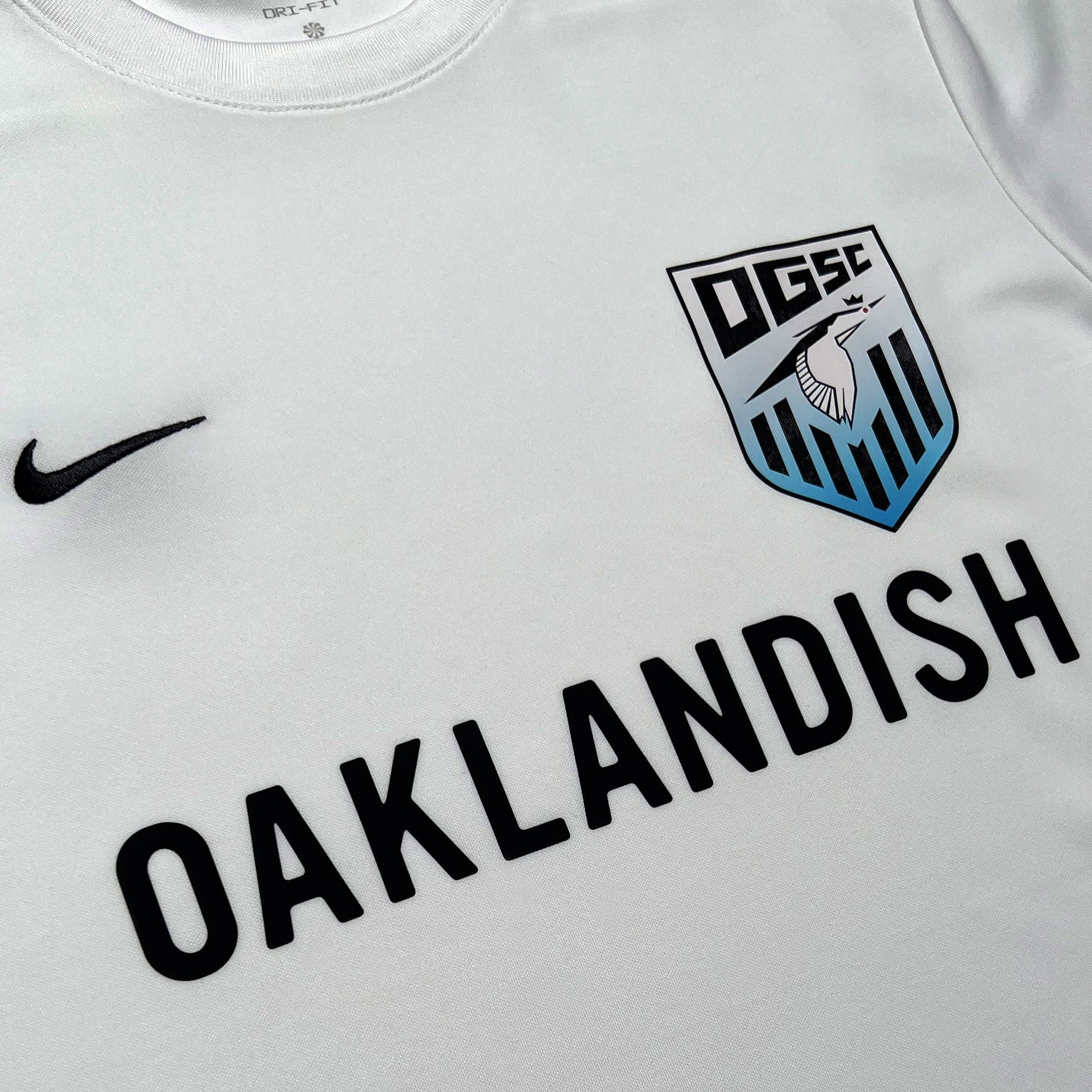 The Oakland Genesis Home White, by Partner, is a white sports jersey featuring the text "DGSC" and an image of a knight's helmet on the left chest area. A black Nike swoosh adorns the right chest, while "OAKLANDISH" is boldly printed in black across the center to celebrate Oakland Genesis youth soccer.