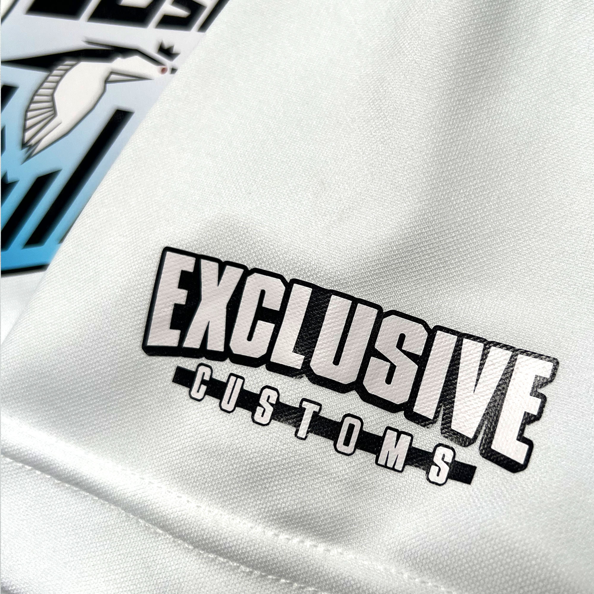 Close-up of a white Oakland Genesis Home White garment by Partner, featuring a bold, black-and-white "Exclusive Customs" logo in an eye-catching blocky font style. A graphic with a bird in flight and stylized text in black, white, and blue appears on the left side. Inspired by the youth soccer nonprofit organization Oakland Genesis.