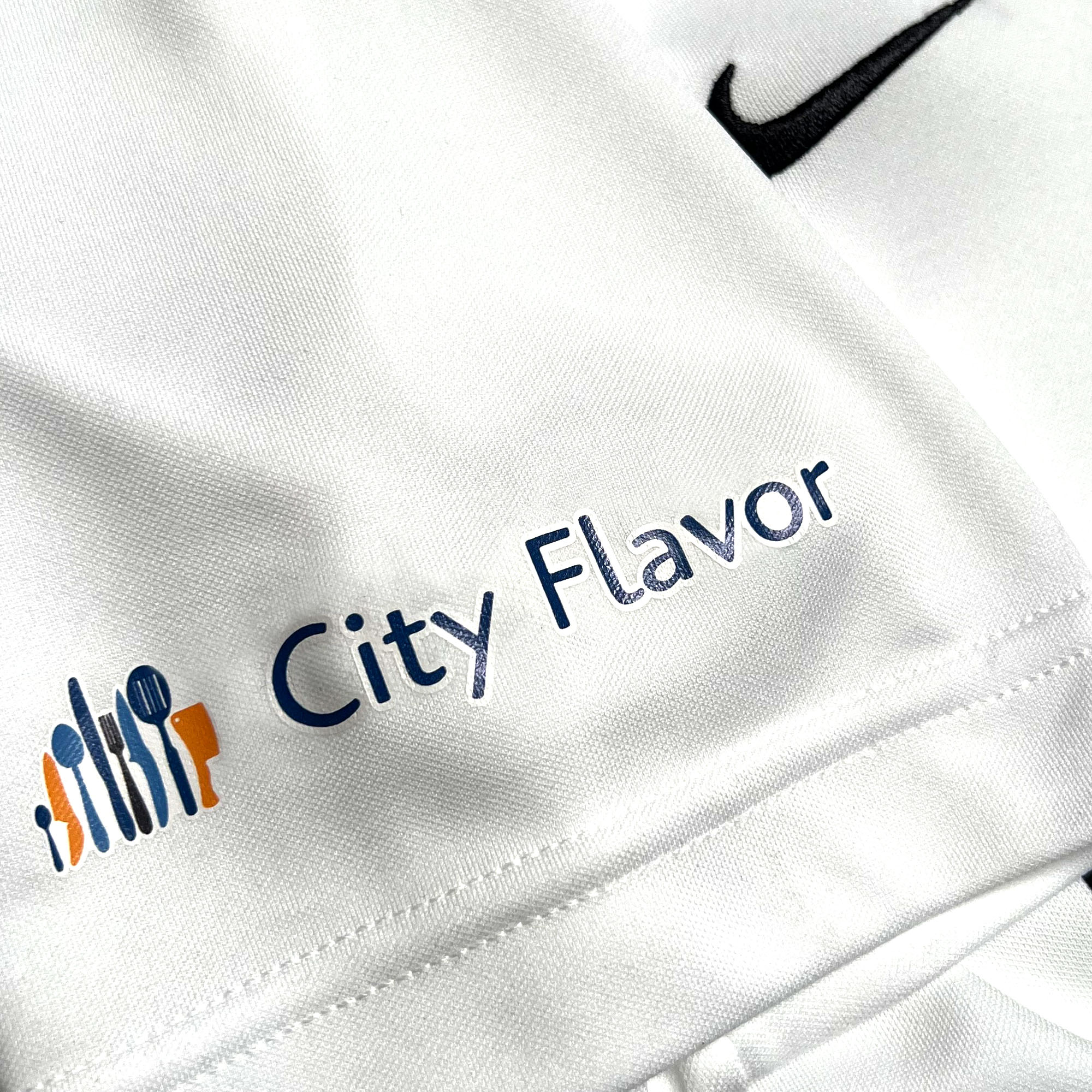 A close-up of the Oakland Genesis Home White fabric shows the text "City Flavor" in blue, along with a logo on the left that features various kitchen utensils—fork, knife, spoon, and spatula—in orange, blue, and green. Additionally, there's an embroidered black Nike swoosh logo at the upper right. Proceeds from this product support Oakland Genesis youth soccer programs through this Partner nonprofit organization initiative.