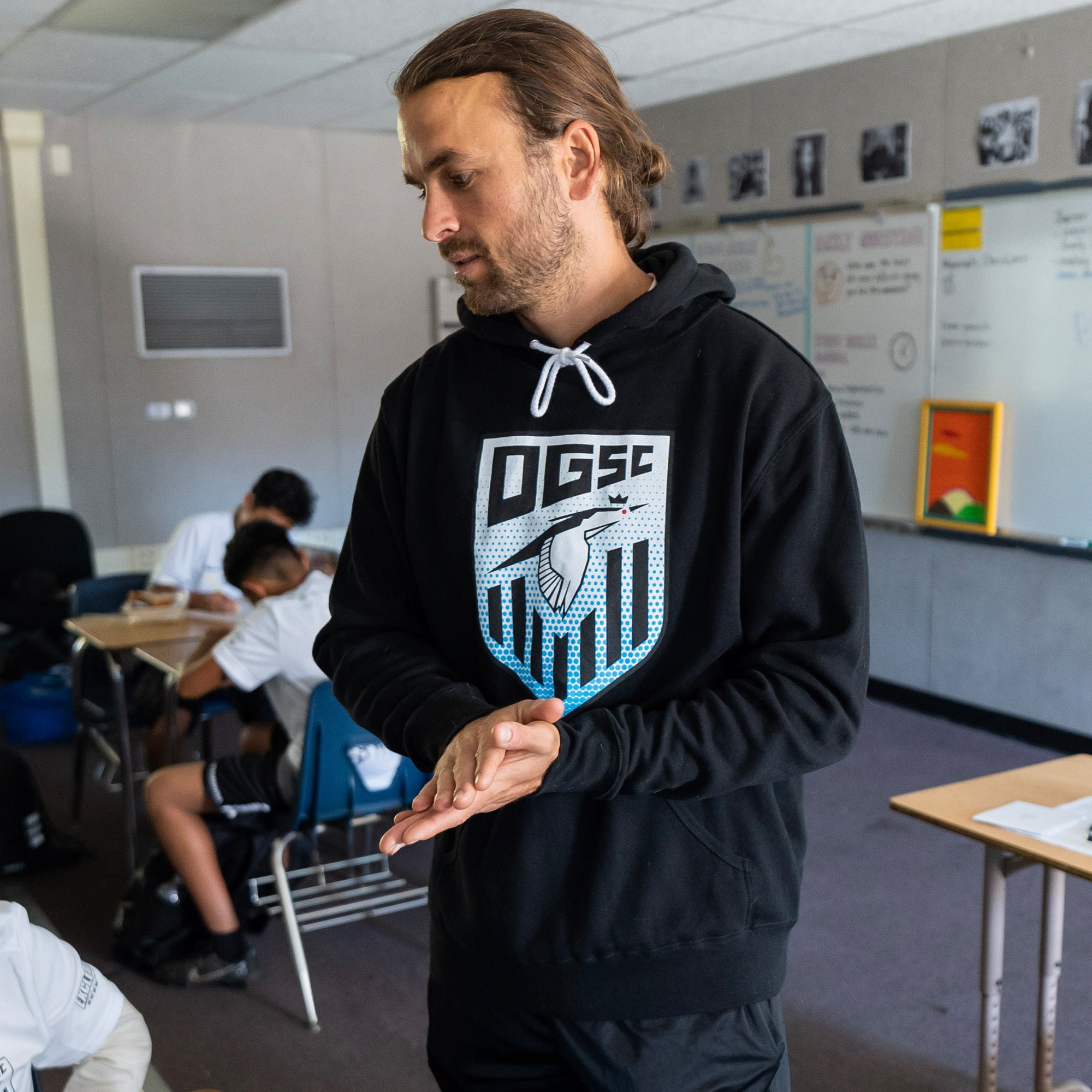 A man with long hair tied back stands in a classroom, wearing a black Oakland Genesis Heron Hoodie by Partner. He clasps his hands while looking down. In the background, students sit on chairs, and a whiteboard with various notes related to youth soccer is visible.