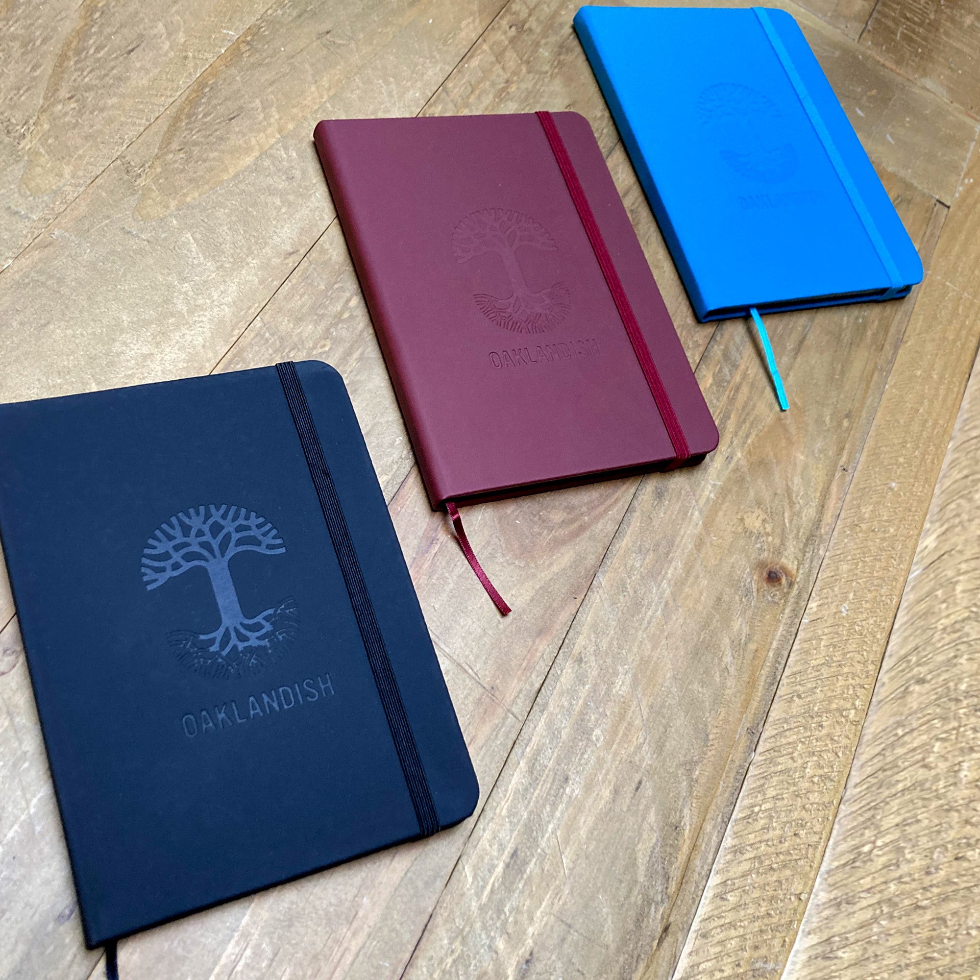 Three Oaklandish Emblem Journals with 80-page lined interiors and elastic straps lie on a wooden surface. The PVC cover journals, available in black, maroon, and bright blue, showcase the debossed Oaklandish tree logo with the word "Oaklandish" below it. The journals are slightly angled and spread out.