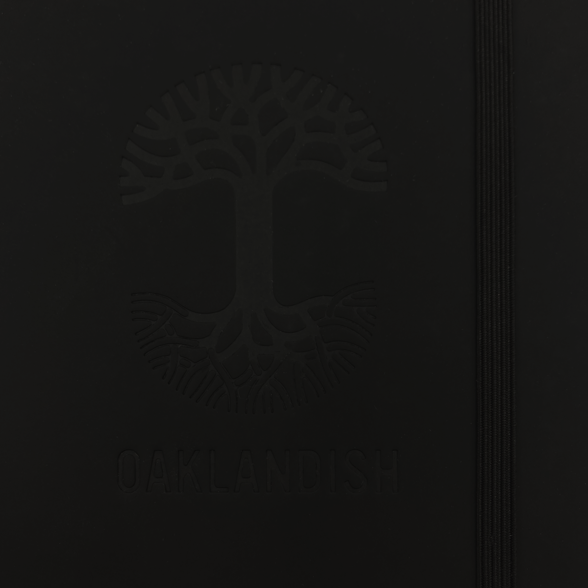 The Oaklandish Emblem Journal, by Oaklandish, features a durable black PVC cover with a debossed Oaklandish tree logo on the front. The logo showcases a broad canopy and intricate root system, with the text "OAKLANDISH" debossed below it. An elastic closure band runs along the right-hand side to keep the journal secure.