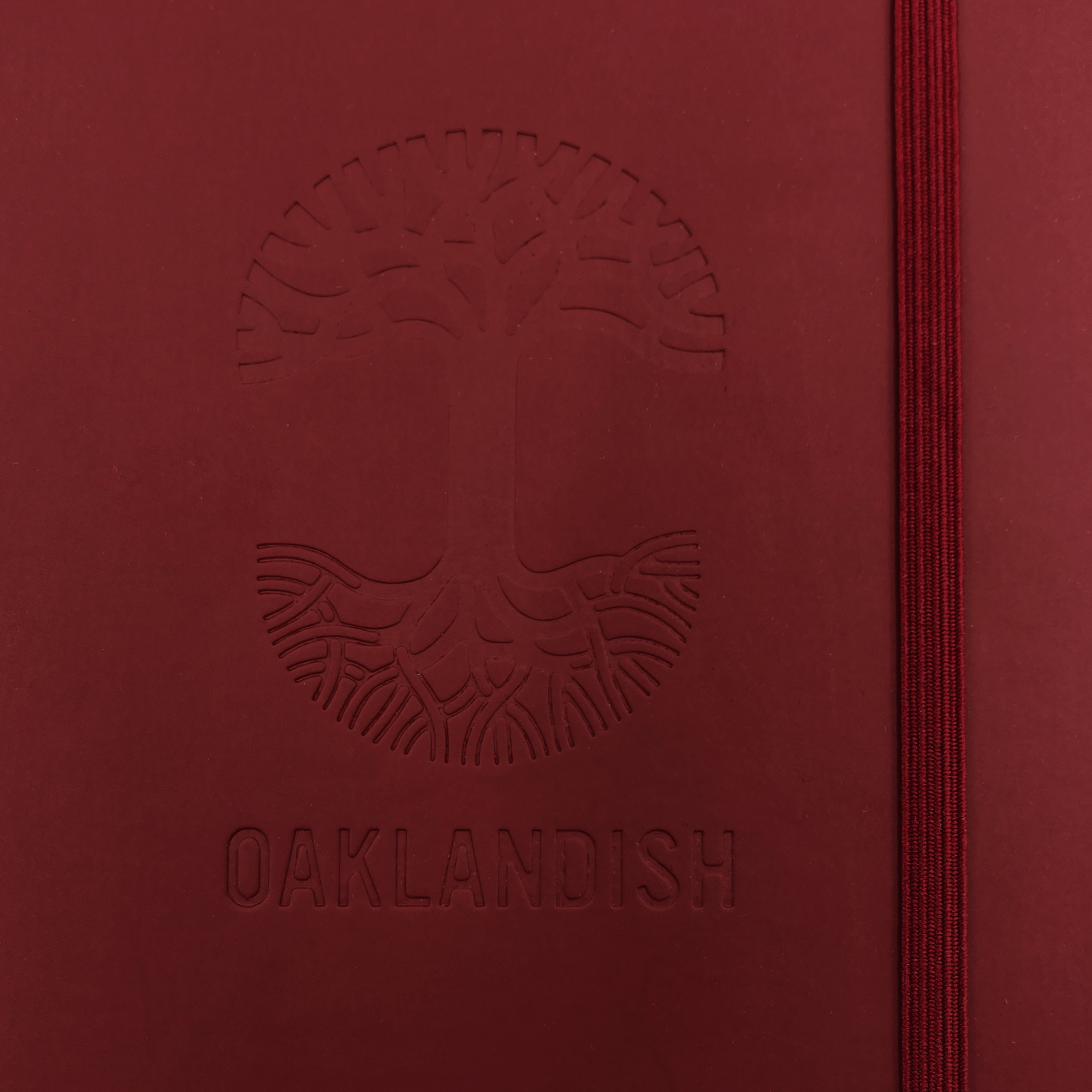 The Oaklandish Emblem Journal, produced by the brand Oaklandish, features a red PVC cover with a debossed tree logo and roots, along with the word "OAKLANDISH" beneath it. This 80-page lined notebook includes a red elastic band closure on the right edge. The design and text are subtly defined through the raised embossing.