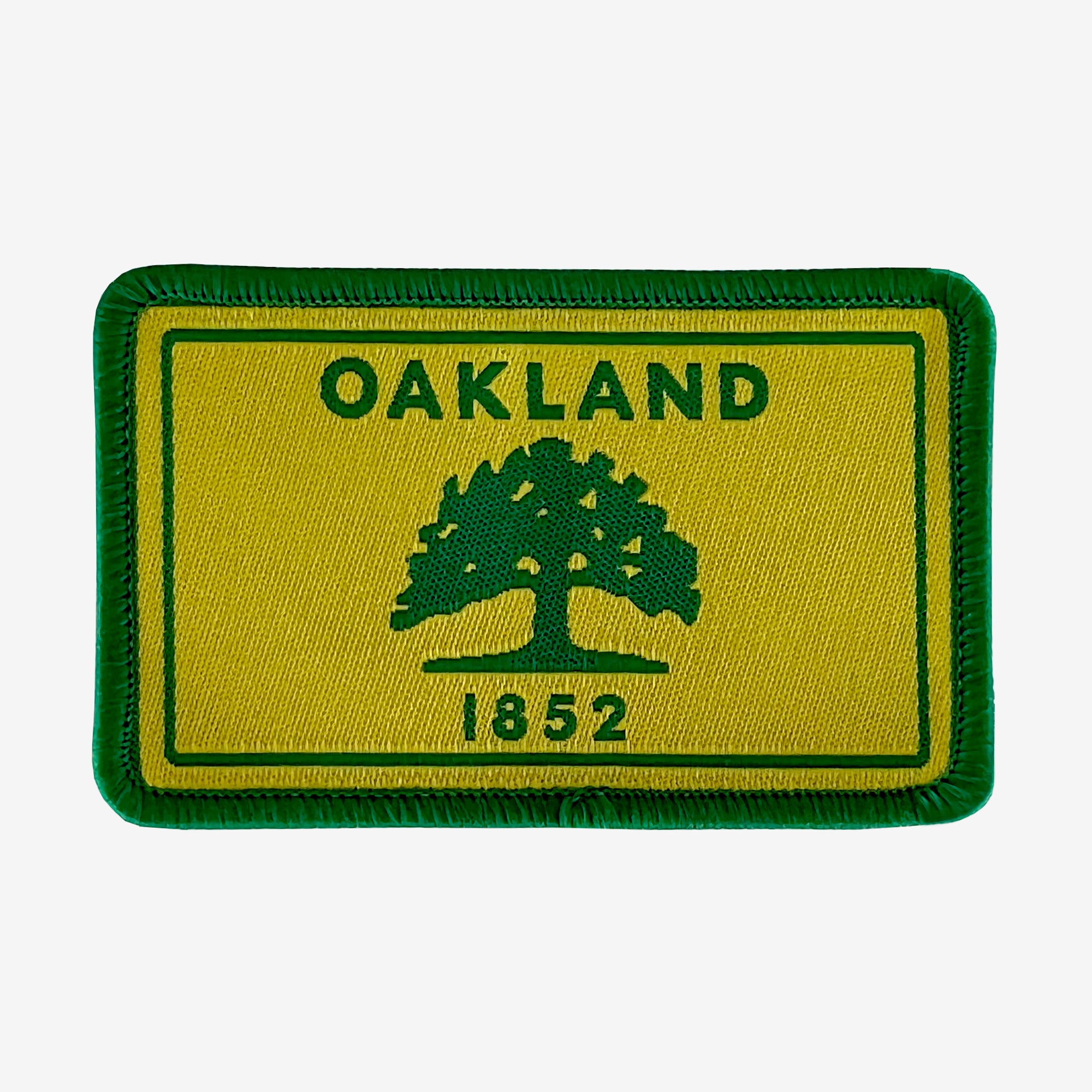 The Oaklandish Oakland Flag Patch is a rectangular, embroidered iron-on patch featuring a green border and a yellow background with a green tree in the center. "OAKLAND" is written in green letters above the tree, and "1852" appears below it. Simply apply with a hot iron for easy attachment.