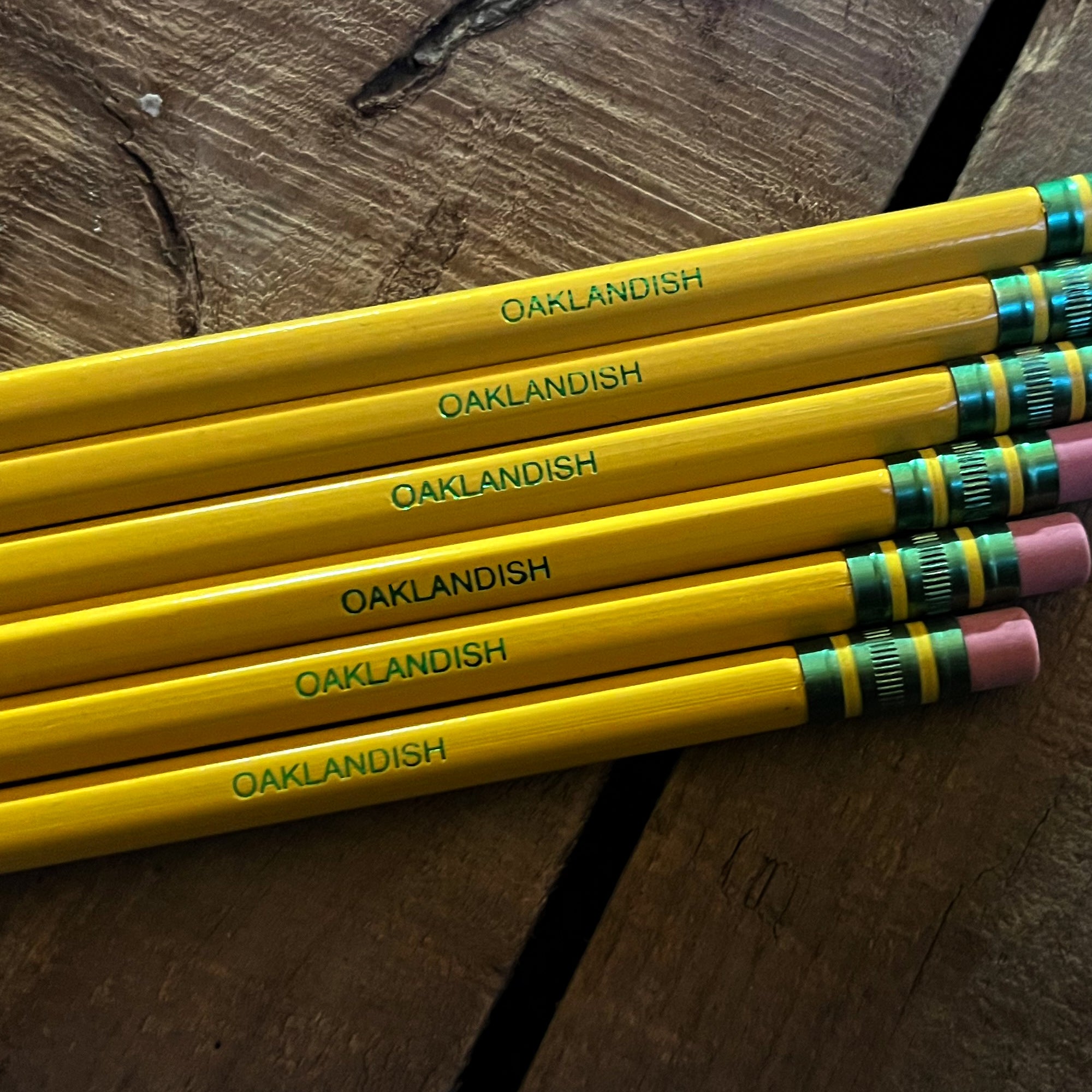 A group of five Oaklandish Pencil Dixon Ticonderoga No. 2 yellow pencils, with "OAKLANDISH" printed in green near the latex-free pink erasers, lies on a wooden surface. Each pencil features a green and gold ferrule. The wood grain of the surface shows visible cracks, casting slight shadows around the pencils.