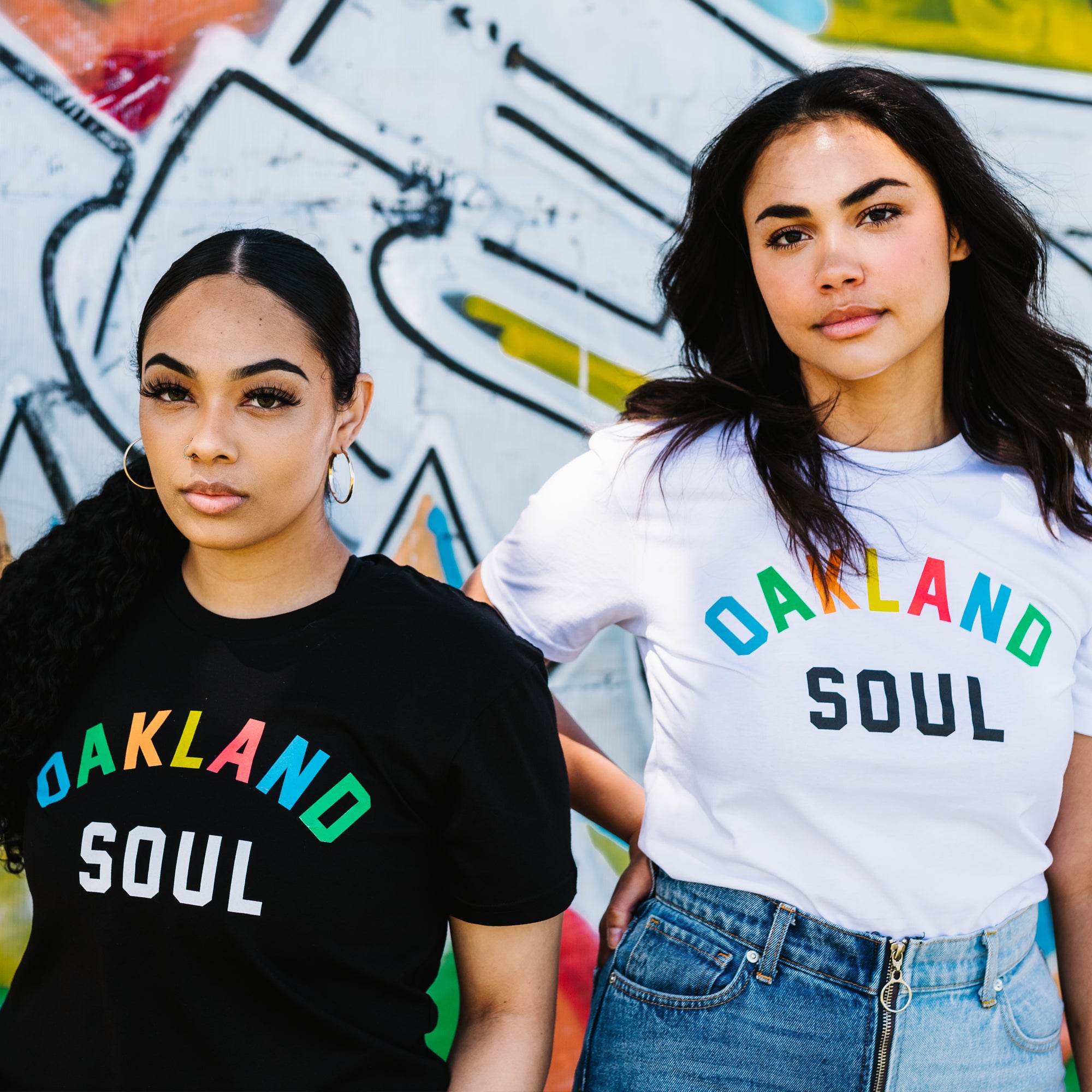 Two women stand in front of a graffiti wall. Both are wearing Oakland Soul Wordmark Tees from Oakland Roots SC, with the phrase "Oakland Soul" displayed in colorful letters. The woman on the left has her hair in a sleek ponytail and accessorizes with large hoop earrings while wearing a black tee. The woman on the right has wavy hair, pairing her white tee with blue jeans.