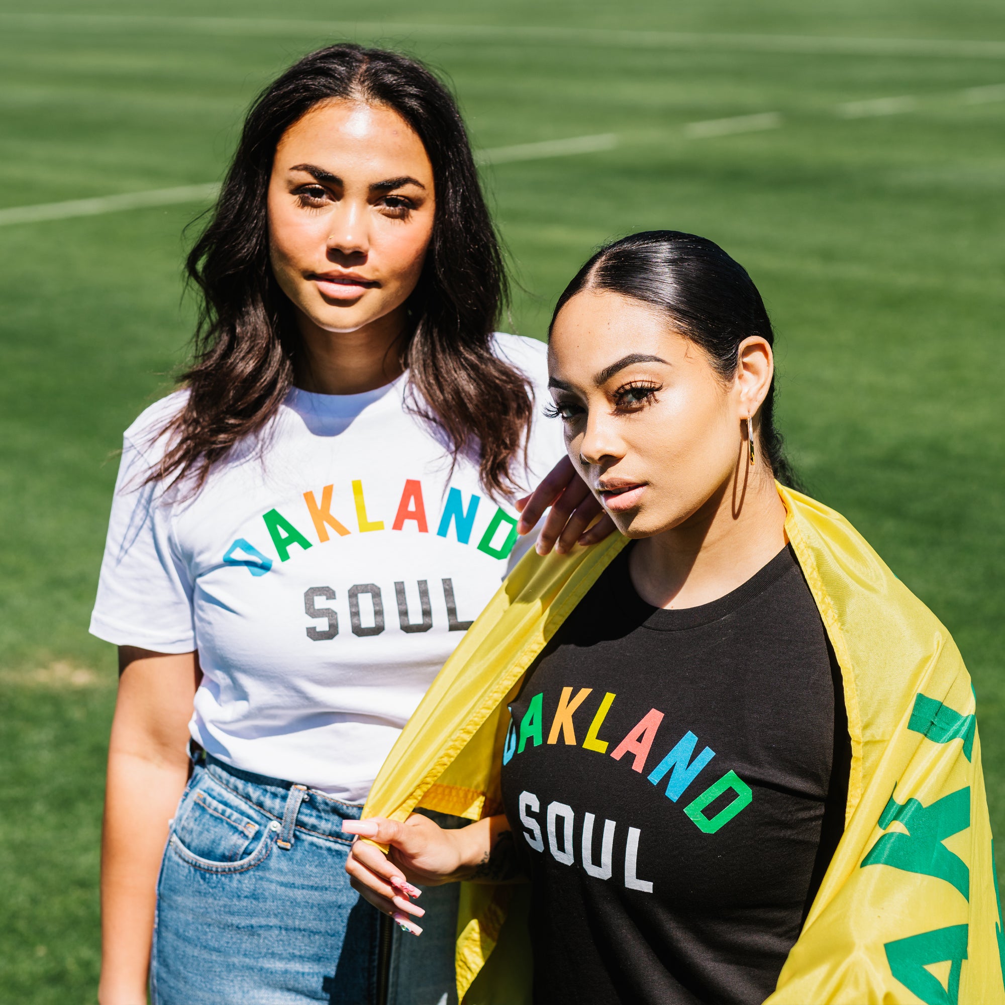 Two women standing on grass wear "Oakland Soul Wordmark Tee" classic fit t-shirts by Oakland Roots SC. One is dressed in a white shirt and blue jeans, while the other pairs her black shirt with a yellow jacket. They strike confident poses, standing closely together against the backdrop of a blurred grassy field.