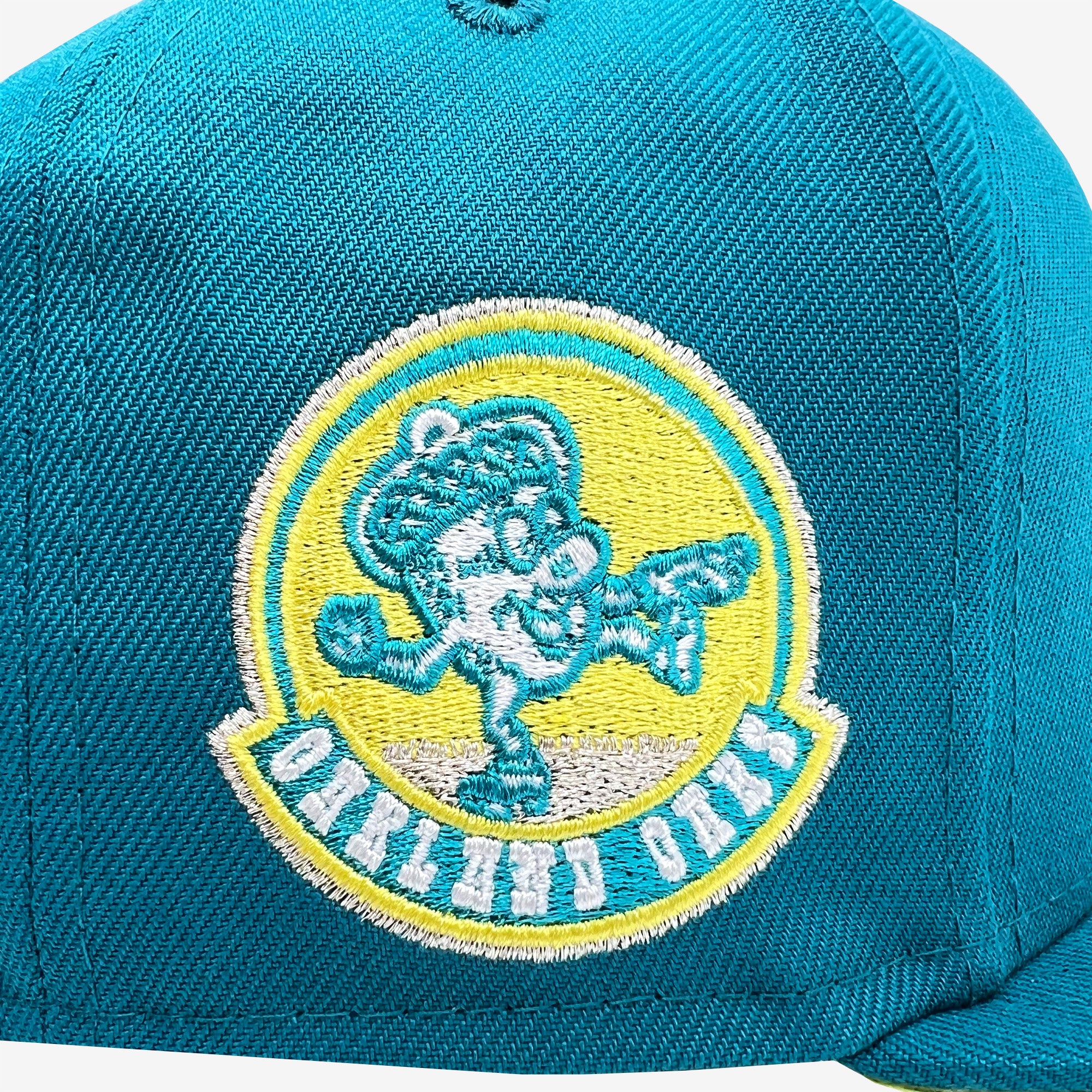 A New Era Oakland Oaks 59FIFTY Fitted Cap from Retail Vendors in teal, featuring a fluorescent yellow embroidered logo of a cheerful cartoon elephant wearing a baseball cap and balancing on one leg with the other leg and trunk raised. The logo is encircled by a yellow border with "OAKLAND OAKS" stitched below.