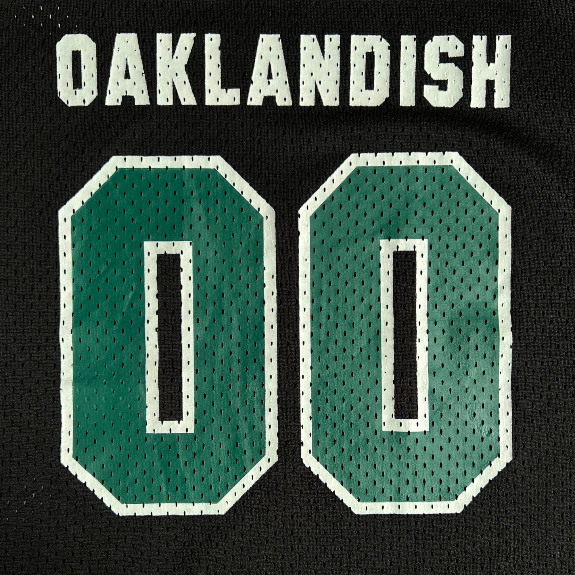 The Official Football Home Jersey by Oaklandish features the word "OAKLANDISH" in white capital letters across the top on a black base. Below, it displays the number "00" in green with white outlines. Made from lightweight mesh fabric, this jersey enhances breathability with small holes visible throughout.