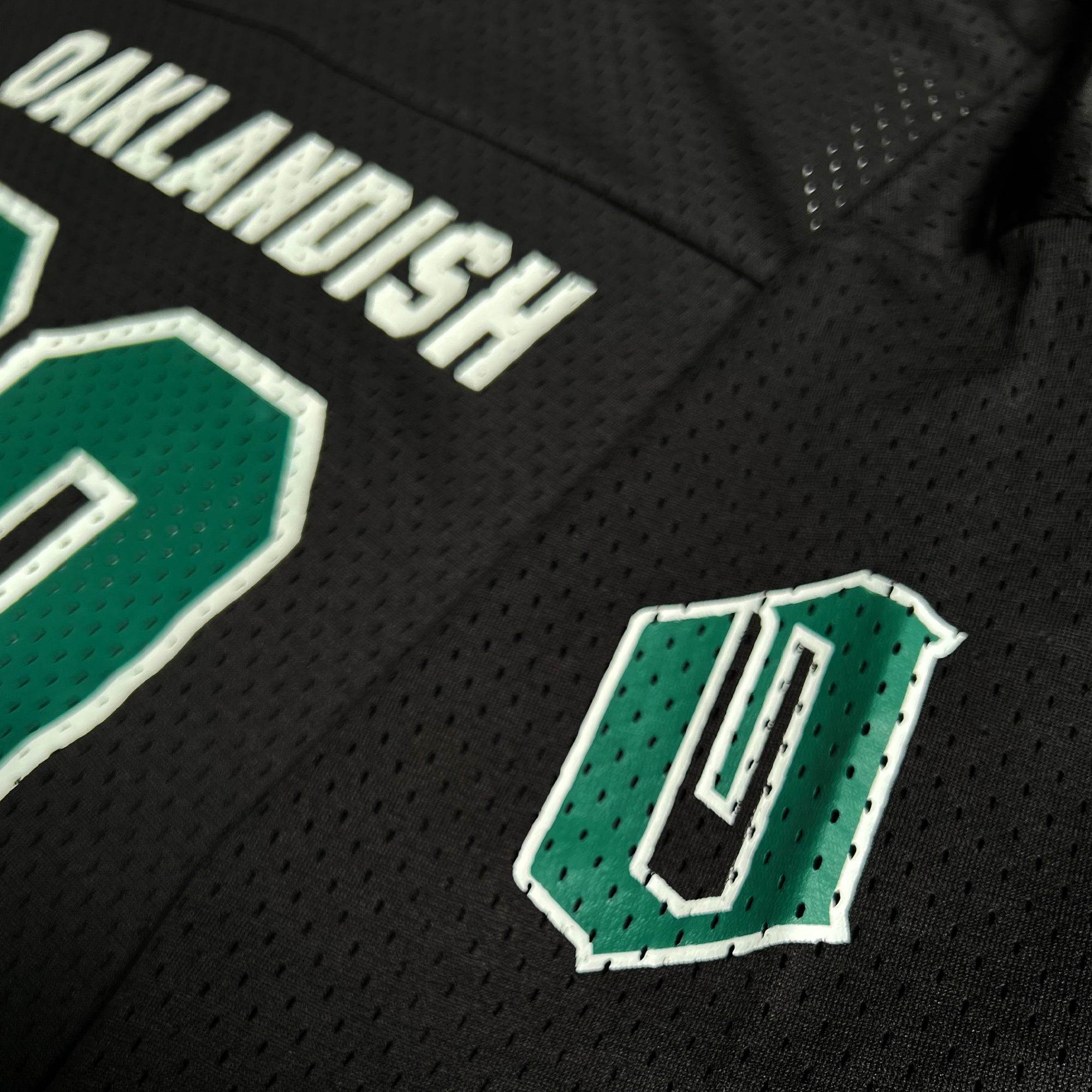 Close-up of the Official Football Home Jersey by Oaklandish, showcasing a sleek black design with white and green detailing. The upper back of this lightweight mesh jersey displays the name "Oaklandish" in bold white letters, with partially visible green and white numbers below it. On the front, a green emblem featuring a stylized "O" outlined in white stitching highlights its breathable construction.