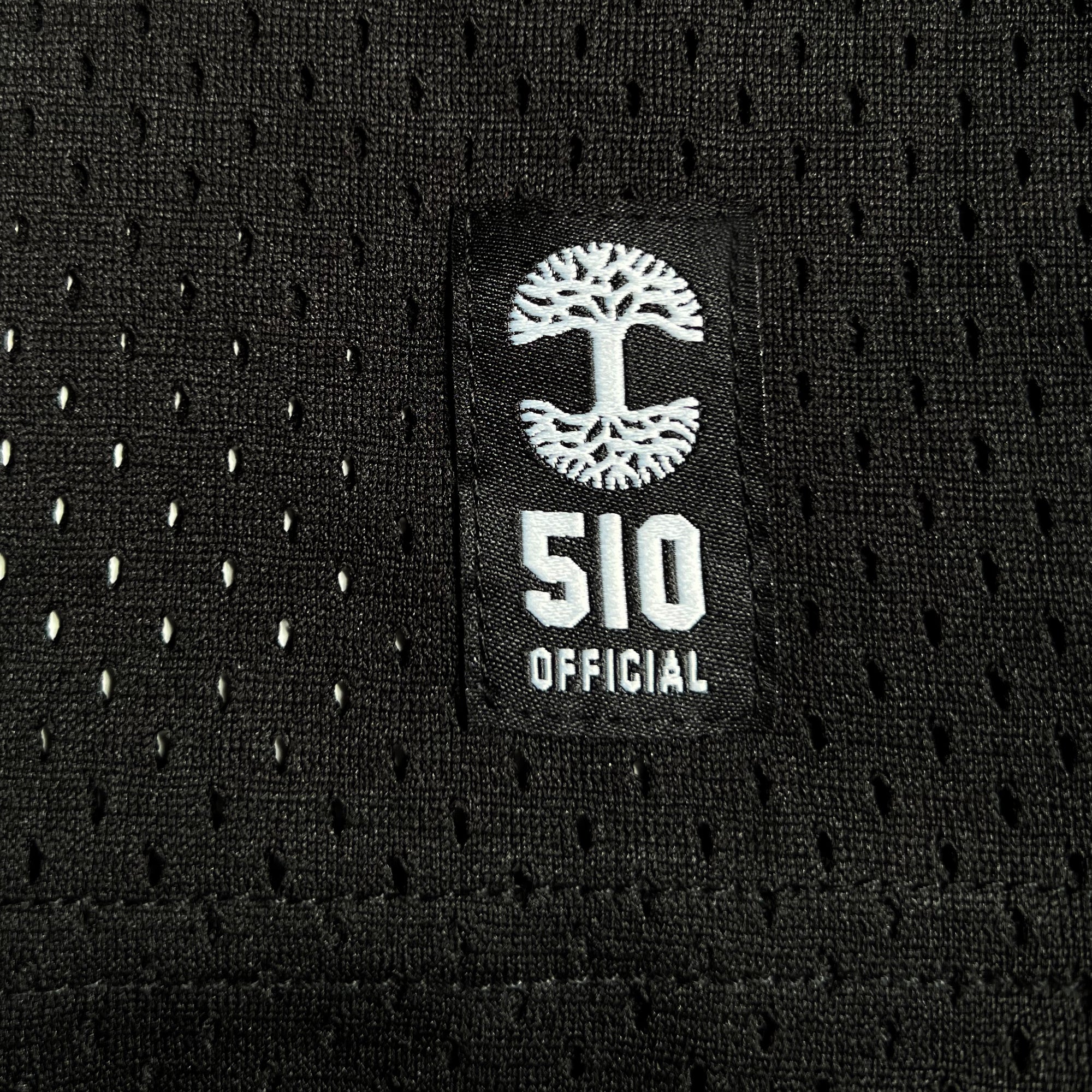 A close-up of a black, perforated athletic fabric reminiscent of the Official Football Home Jersey by Oaklandish features a rectangular logo. The logo displays a stylized white tree above the text "510 OFFICIAL." The tree has a symmetrical, spreading canopy and root structure, creating a mirror image effect. The lightweight mesh enhances breathability with numerous small holes.