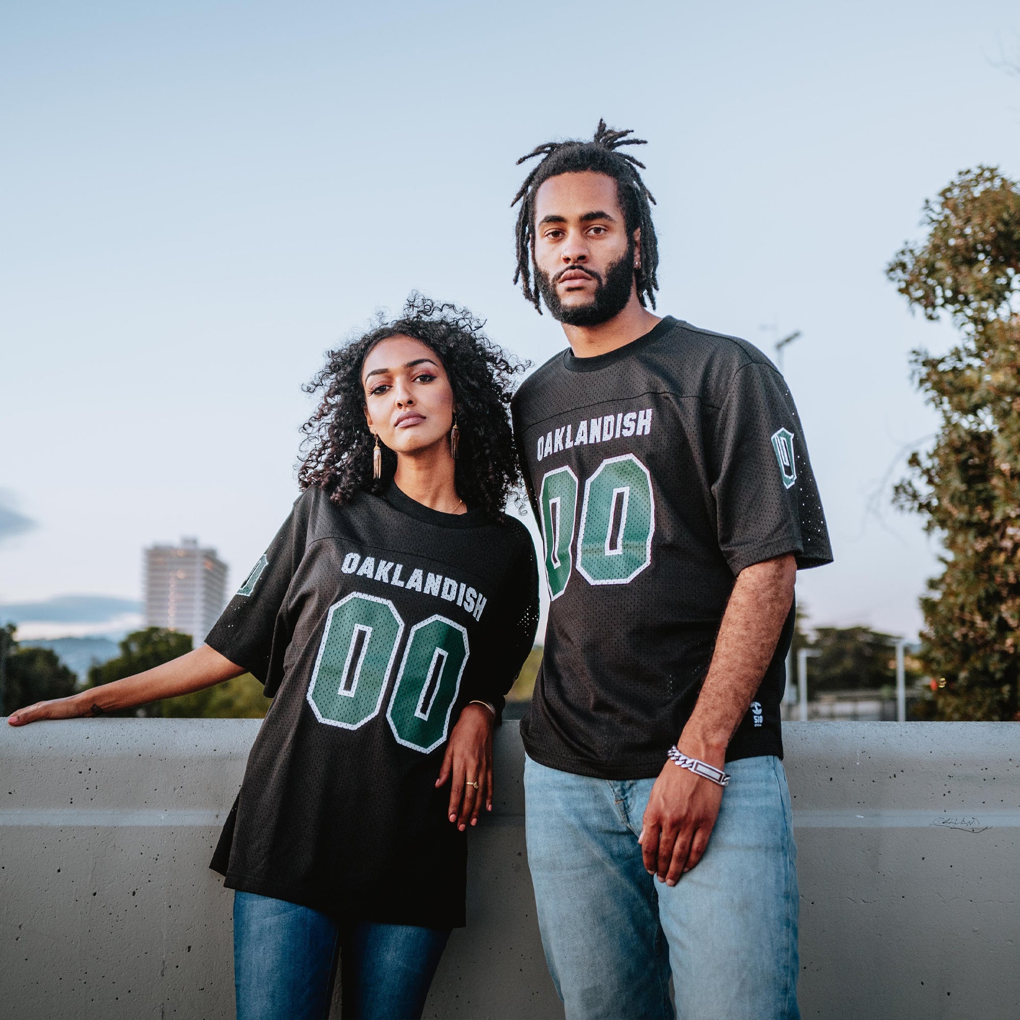 A woman and a man stand side by side outdoors on a concrete structure, both donning matching black "Official Football Home Jerseys" by Oaklandish, made from lightweight mesh. The woman, with curly hair, rests her left arm on the divider. The man has dreadlocks and a beard, with trees and buildings in the background.