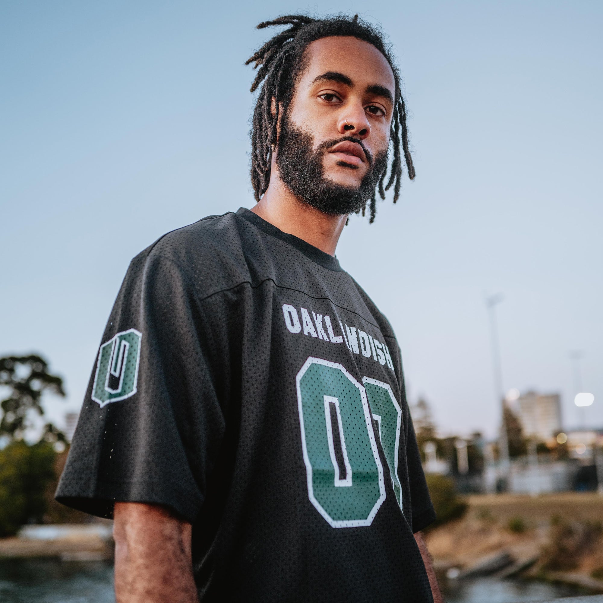 A man with a beard and dreadlocks stands outdoors, wearing the Official Football Home Jersey from Oaklandish. The black jersey is emblazoned with the number 01 and the word "OAKLANDISH." Its lightweight mesh design ensures breathability as he gazes confidently at the camera. In the background, a park area with trees, a water body, and a city skyline are visible under a clear sky.