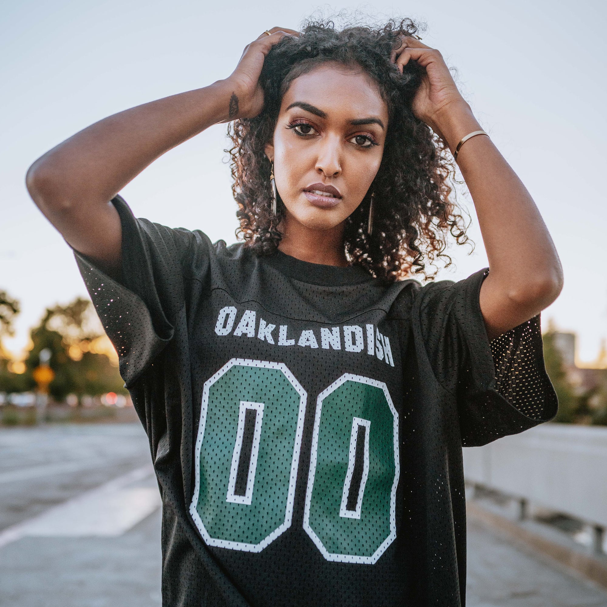 A person with curly hair stands outdoors, looking at the camera. They are wearing the Official Football Home Jersey by Oaklandish, a black lightweight mesh football jersey with "Oaklandish" and the number "00" on the front. The background features trees and a walkway, suggesting a park or urban setting. The overall mood is casual and relaxed.