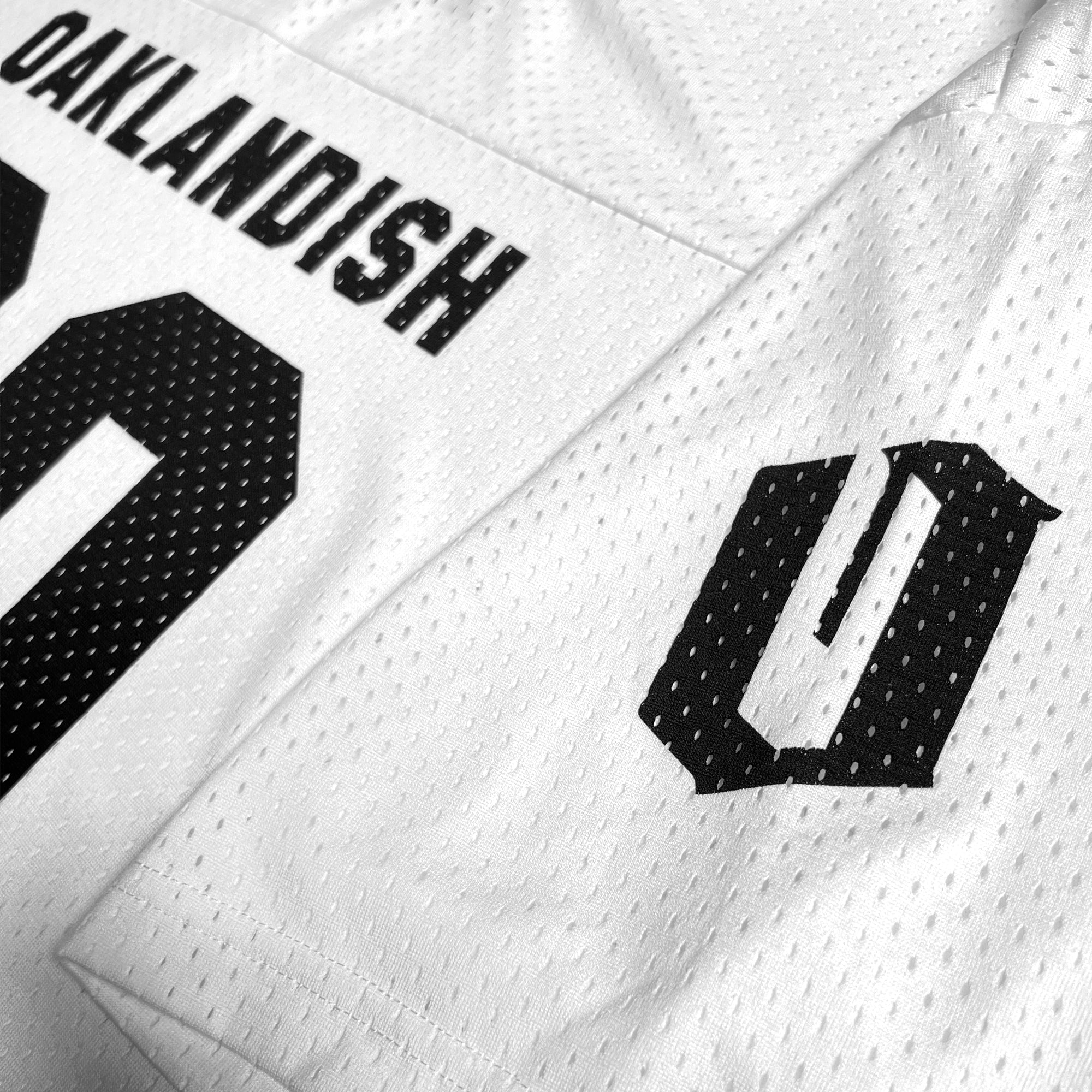 A close-up photo of the Official Football Away Jersey from Oaklandish reveals its white lightweight mesh material, featuring the word "OAKLANDISH" and the number "0" in perforated black letters. An adjacent part of the jersey displays a stylized "O" with a vertical line through it, also in black.