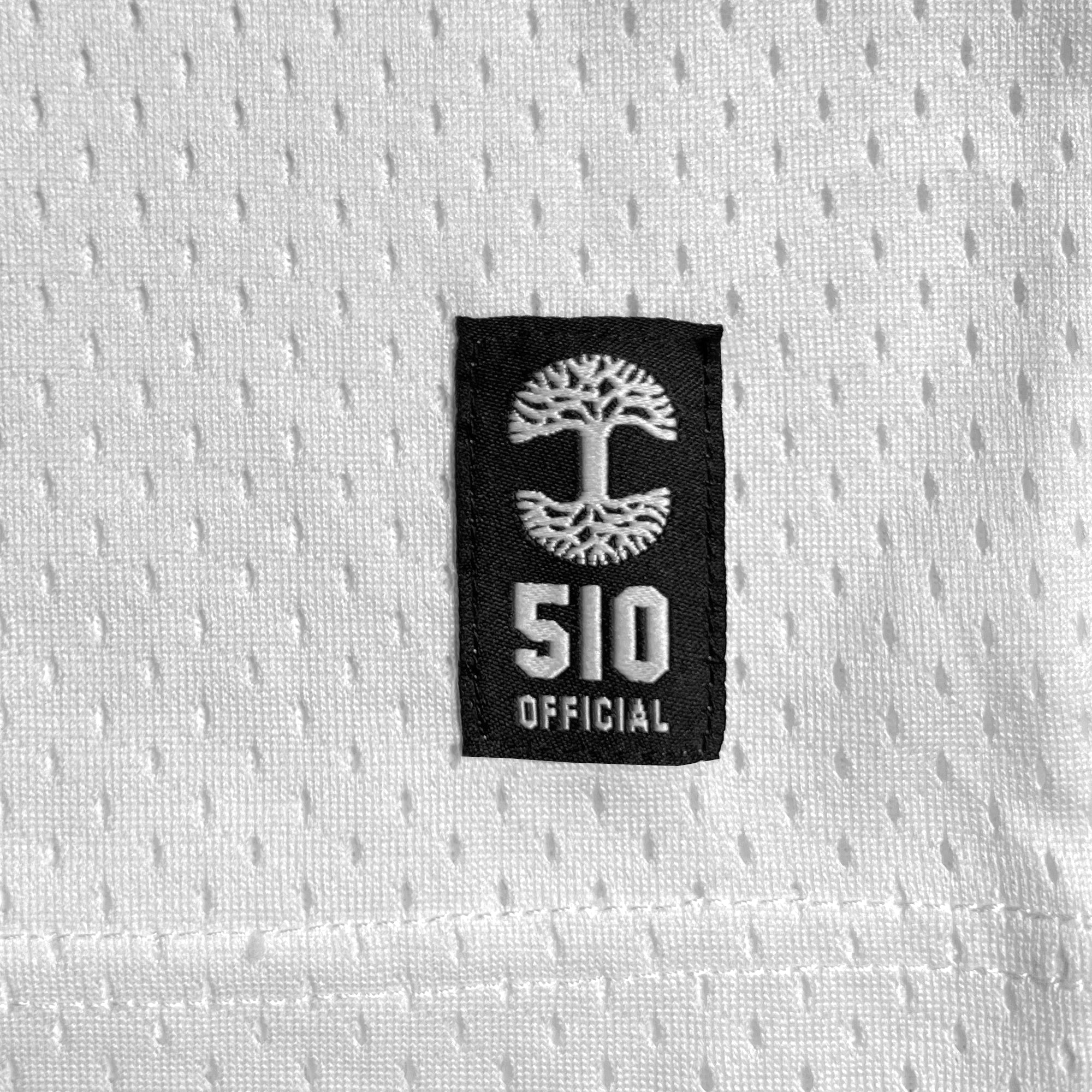 Close-up of Oaklandish's Official Football Away Jersey, showcasing white fabric with a textured, perforated pattern akin to lightweight mesh material. Attached to the jersey is a black rectangular patch featuring Oaklandish's iconic white tree symbol and the text "510 OFFICIAL" in white. The patch distinctly contrasts with the uniform white background.