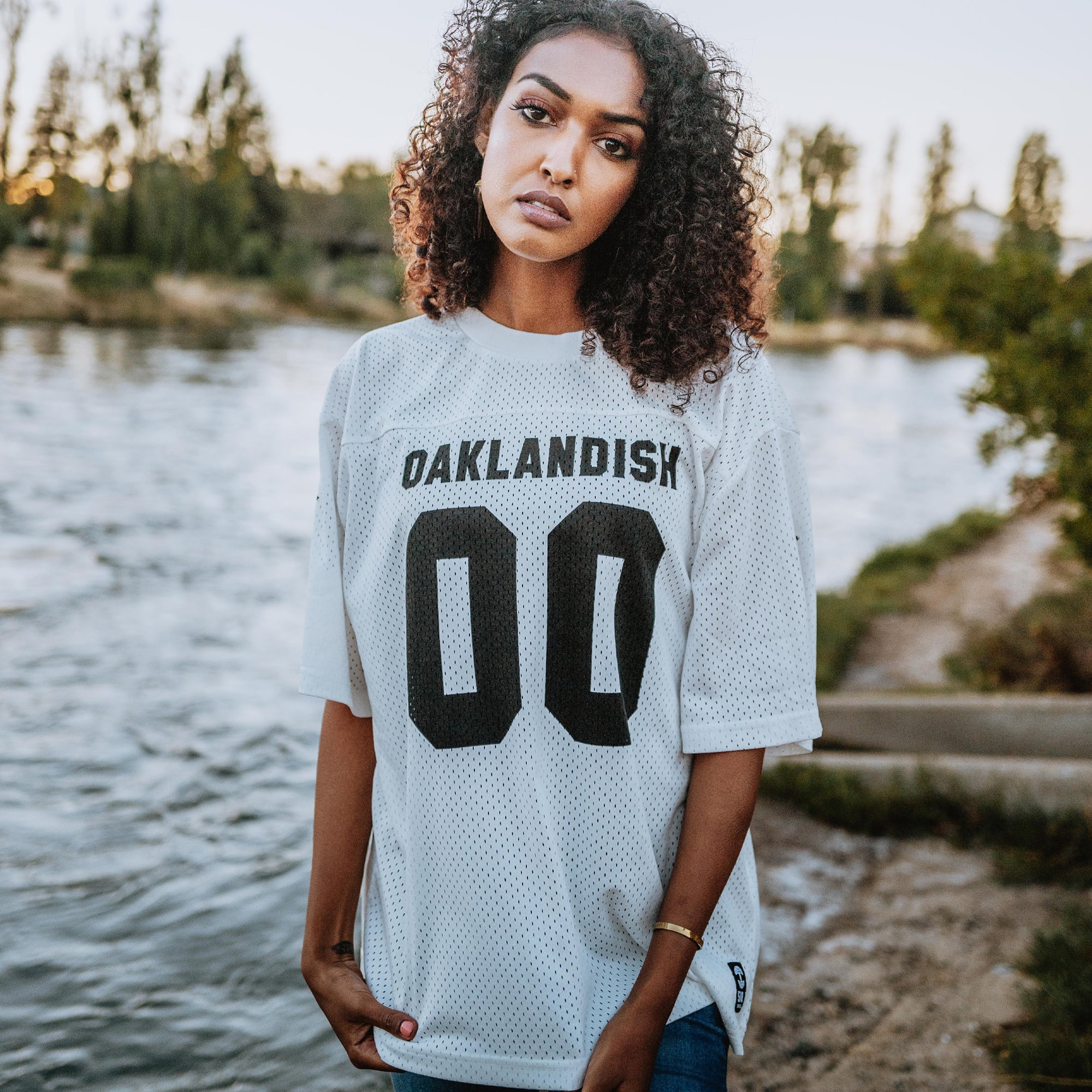 A woman with curly hair, wearing a white Official Football Away Jersey from Oaklandish made from lightweight mesh material and featuring "OAKLANDISH 00" on the front, stands near a body of water. She gazes at the camera with a neutral expression. Trees and a grassy shore are visible in the background. The sky is clear, indicating it is likely late afternoon or early evening.