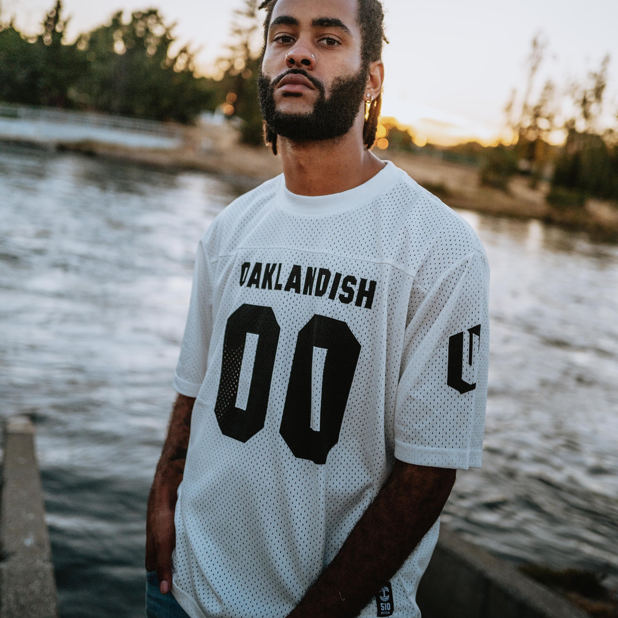 A man with a beard and dreadlocks stands by a river, wearing the Official Football Away Jersey from Oaklandish with "OAKLANDISH 00" printed on it. Trees and a sunset can be seen in the background. His expression is serious as he gazes at the camera.