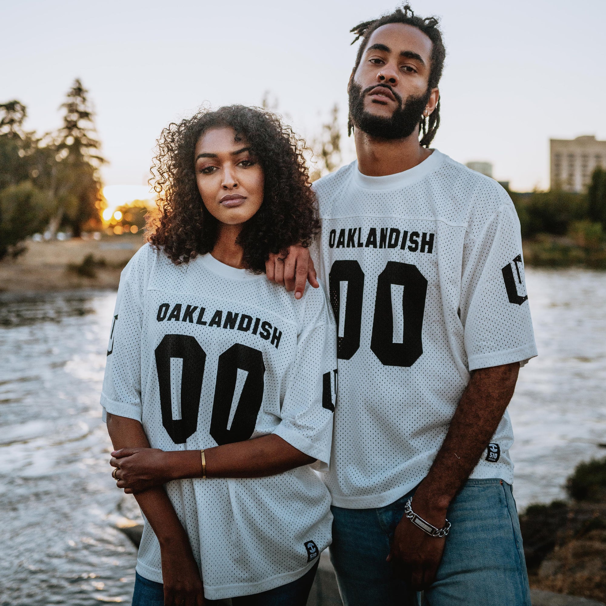 A man with dreadlocks, a beard, and darker jeans stands side by side with a woman who has curly hair and light jeans near a body of water at sunset. Both are wearing Oaklandish's Official Football Away Jerseys, featuring a white, lightweight mesh design with "OAKLANDISH 00" emblazoned on them. Trees and buildings create the backdrop.