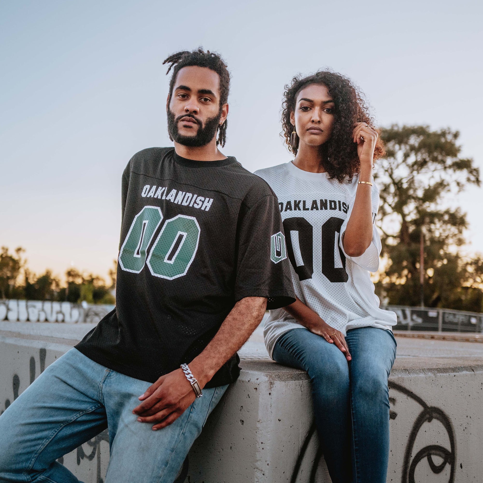 A man and a woman are sitting on a graffiti-adorned concrete ledge during sunset. Both wear Oaklandish Official Football Away Jerseys made from lightweight mesh material, with the man in black and the woman in white. They have naturally curly hair and are casually posed, gazing at the camera. Trees and a clear sky provide the backdrop.