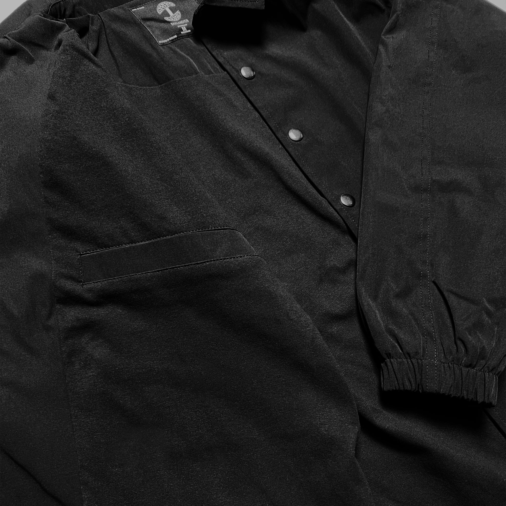 A close-up image of the Oaklandish Premium Coaches Jacket reveals a black garment with a long sleeve that includes gathered elastic at the cuff and a partially visible pocket on the left chest area. The front showcases a button-down design with three visible buttons, and the fabric has a slight sheen, indicative of its premium quality in men's sizing.