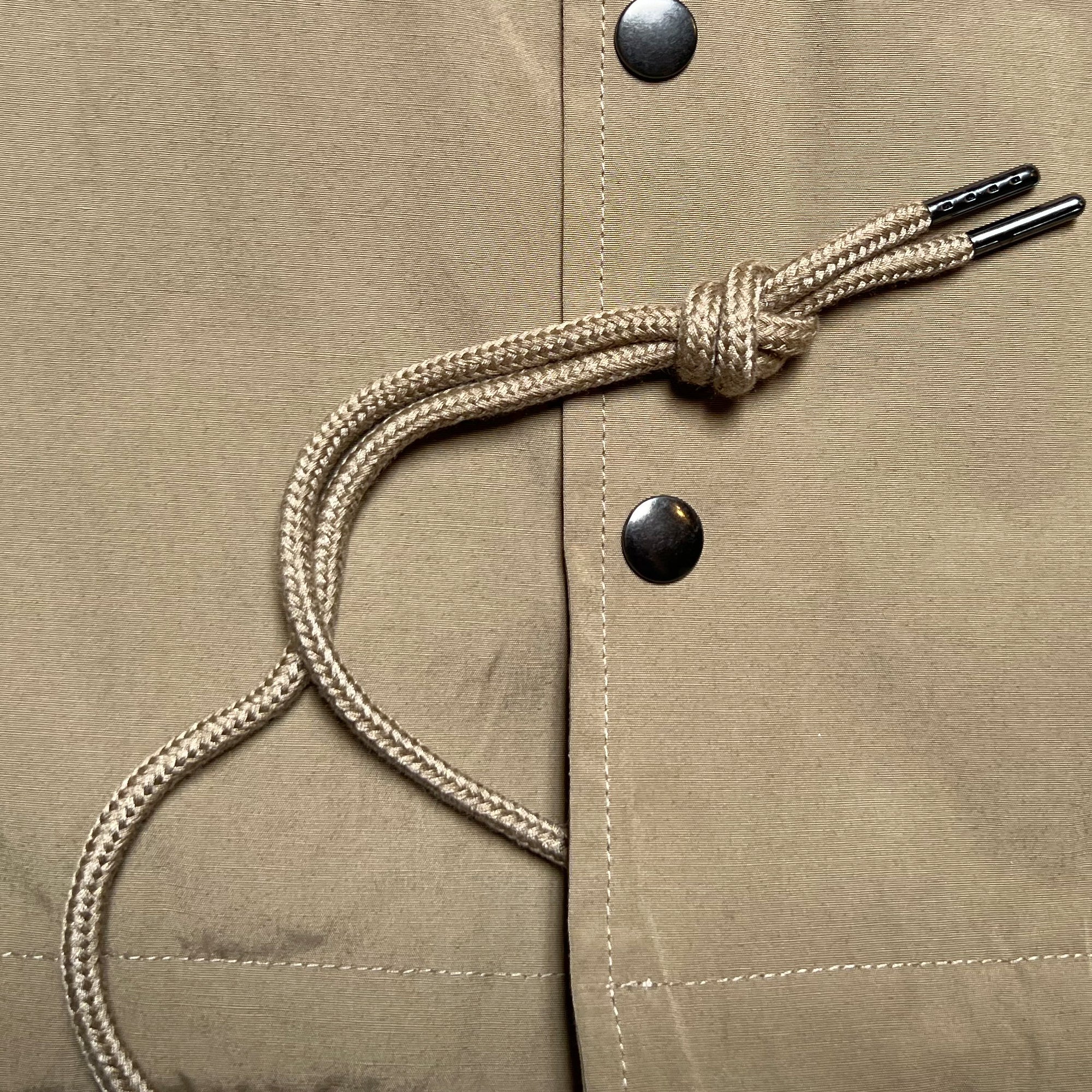 A close-up of the tan, water-resistant fabric on the Oaklandish Premium Coaches Jacket, showcasing metal snap buttons and a beige drawstring tied in a knot with metal-tipped ends. The visible stitching on the fabric forms a neat, clean edge. The image highlights the fine details of this premium men's jacket.