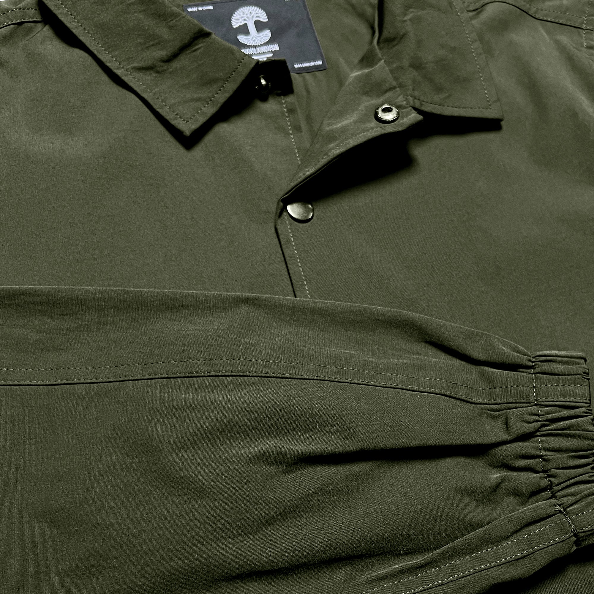 Close-up of the Oaklandish Premium Coaches Jacket that resembles premium outerwear. The smooth, dark green fabric has a subtle sheen under the lighting, and silver snap buttons enhance its style. The gathered sleeve with elastic is prominently displayed in the foreground, alongside a partially visible label with a mushroom icon.