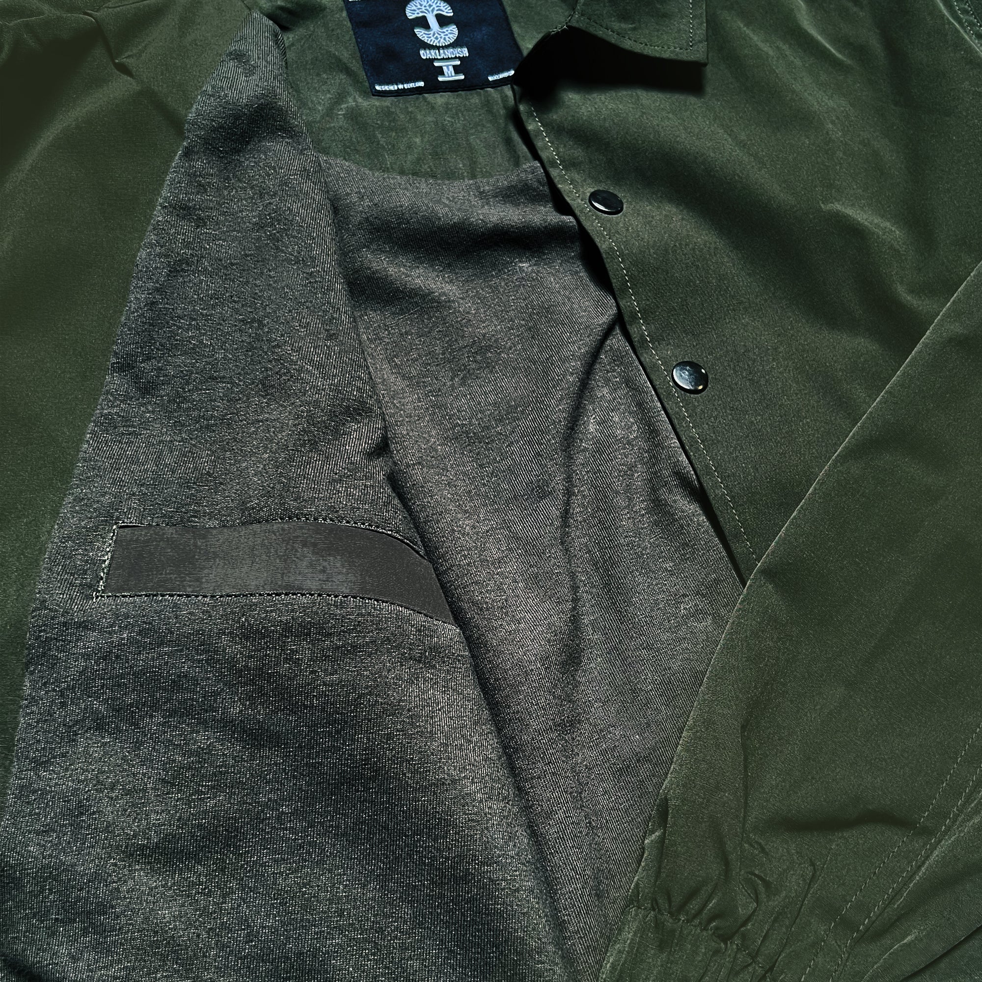 A close-up view of the Oaklandish Premium Coaches Jacket reveals its smooth, slightly shiny outer fabric. The olive green outerwear boasts a soft, darker-shaded inner lining and features black snap buttons. There's also a visible inner pocket on the left side. This jacket is both stylish and water-resistant.