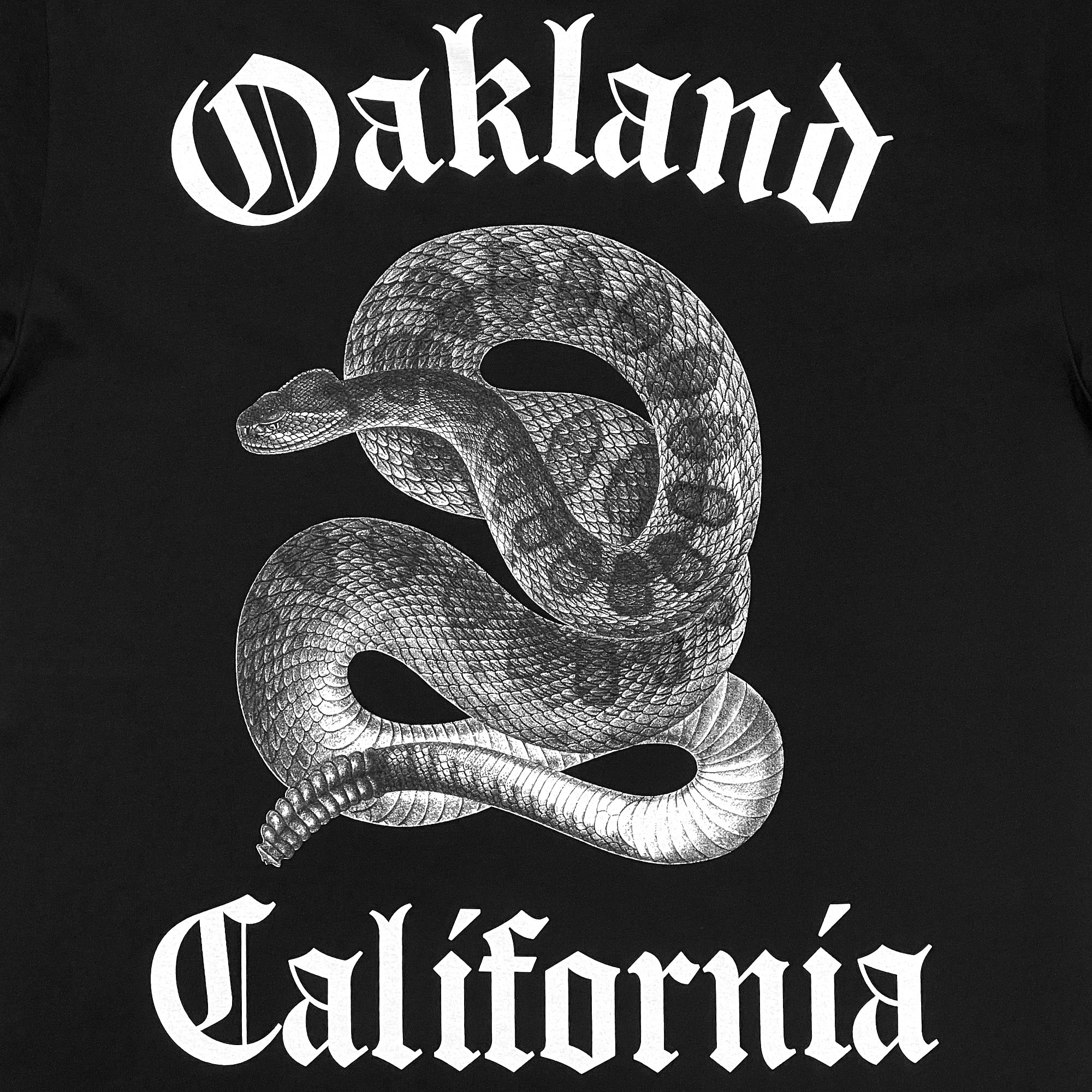 The Sistrurus Tee by Oaklandish features a detailed white illustration of a coiled, venomous snake at its center on a classic fit black t-shirt. The word "Oakland" is boldly displayed above the snake and "California" below it, both in Gothic-style font. Made from 100% cotton for lasting comfort.