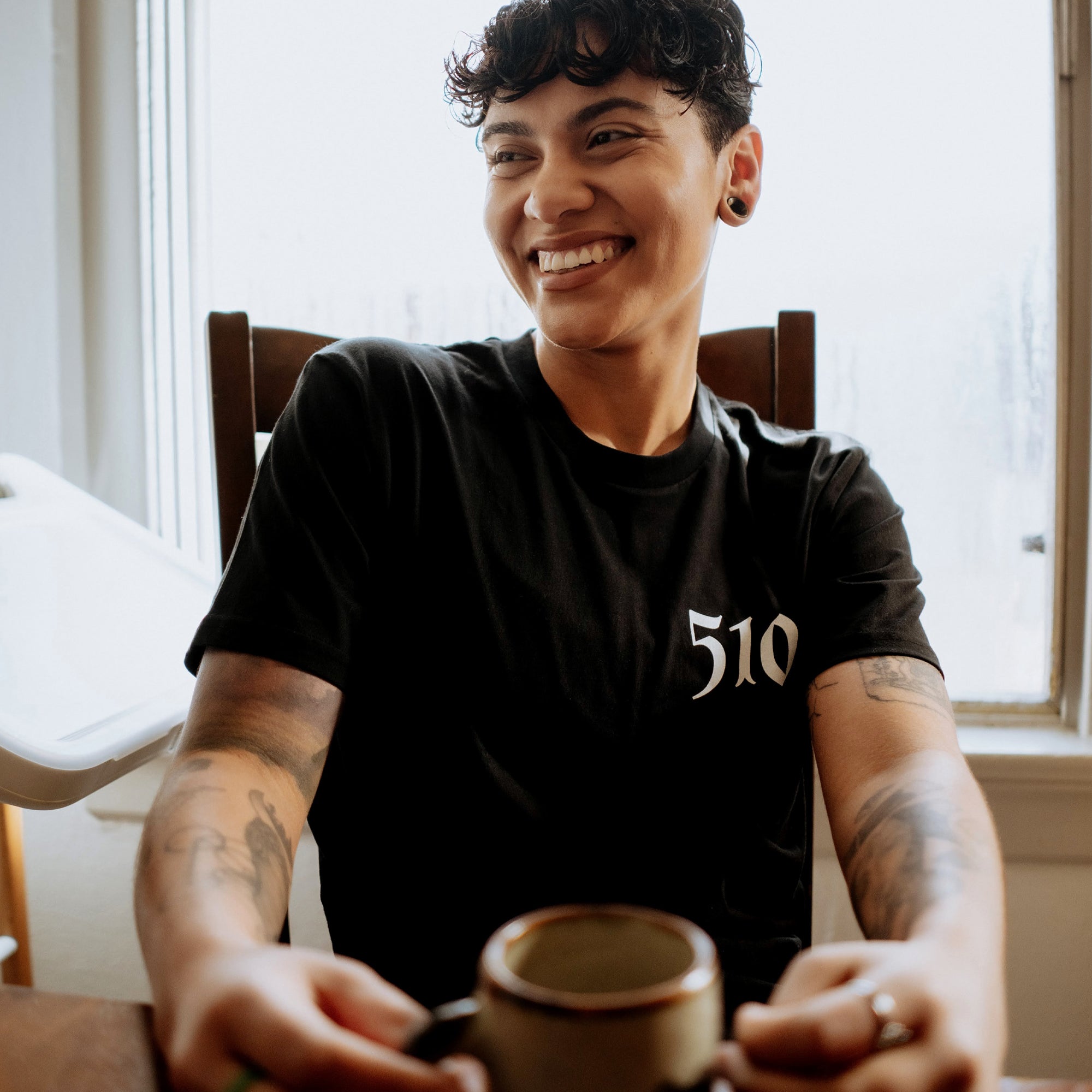 A person with short, curly hair and tattoos on their arms is seated and smiling while holding a mug. They are wearing a black classic fit Sistrurus Tee from Oaklandish with "510" written on it. The background features a window with natural light streaming in, illuminating the room.