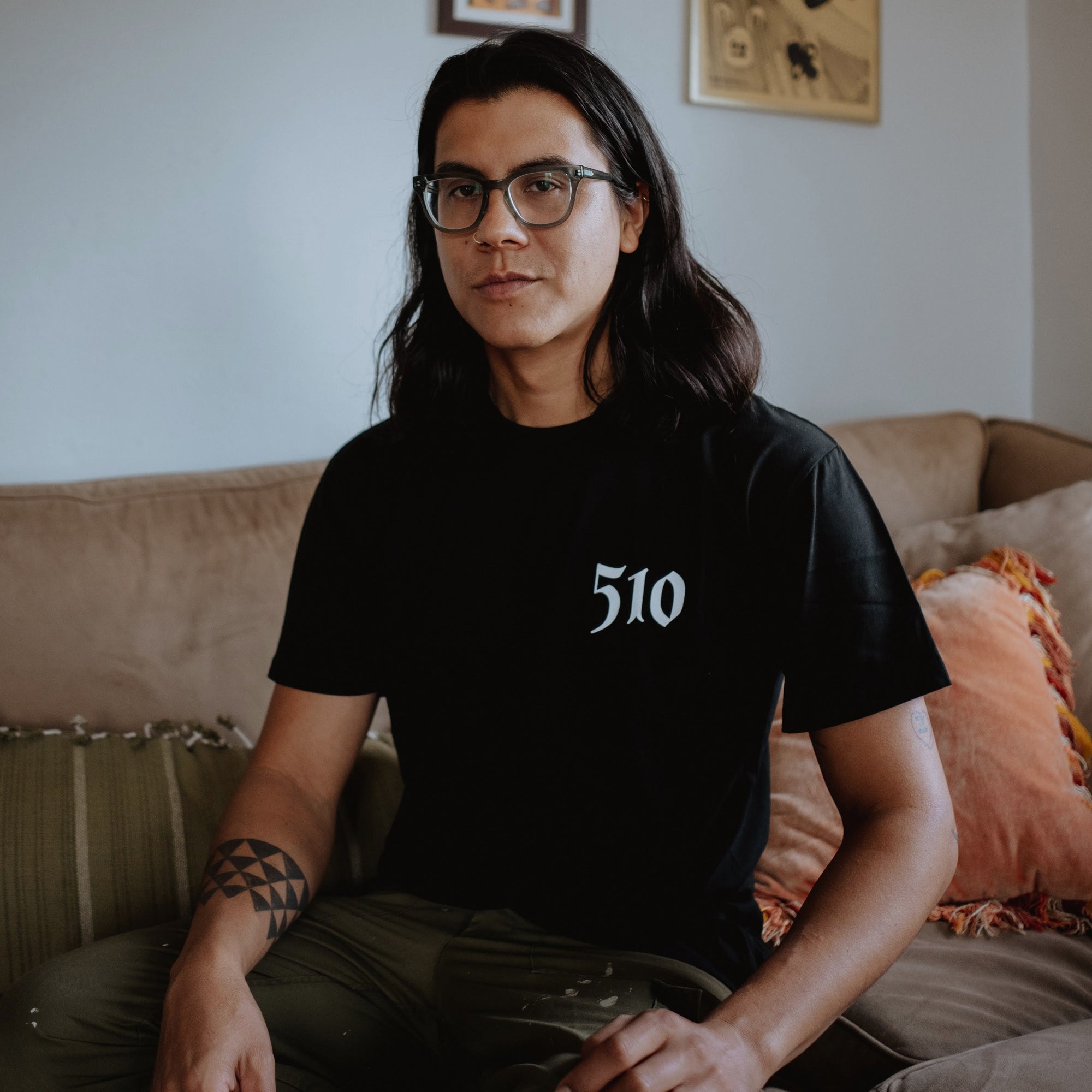 A person with long dark hair and glasses is sitting on a beige couch. They are wearing the Oaklandish Sistrurus Tee, a classic fit t-shirt made of 100% cotton with the number "510" printed on it in white. Their left arm has a geometric tattoo. In the background, there are decorative pillows and two framed pictures on the wall.