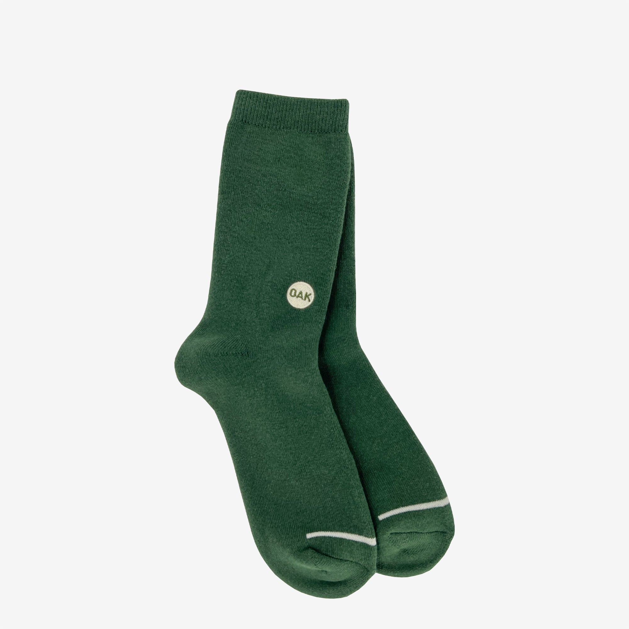 A pair of Oaklandish Plush Oak Socks in dark green is showcased against a plain white background. Each sock is adorned with a small circular Oak logo marked with the letters "QAK" near the top outer side. These plush socks feature a subtle white outline at the toe area and have a ribbed cuff at the top, ensuring happy feet all day long.