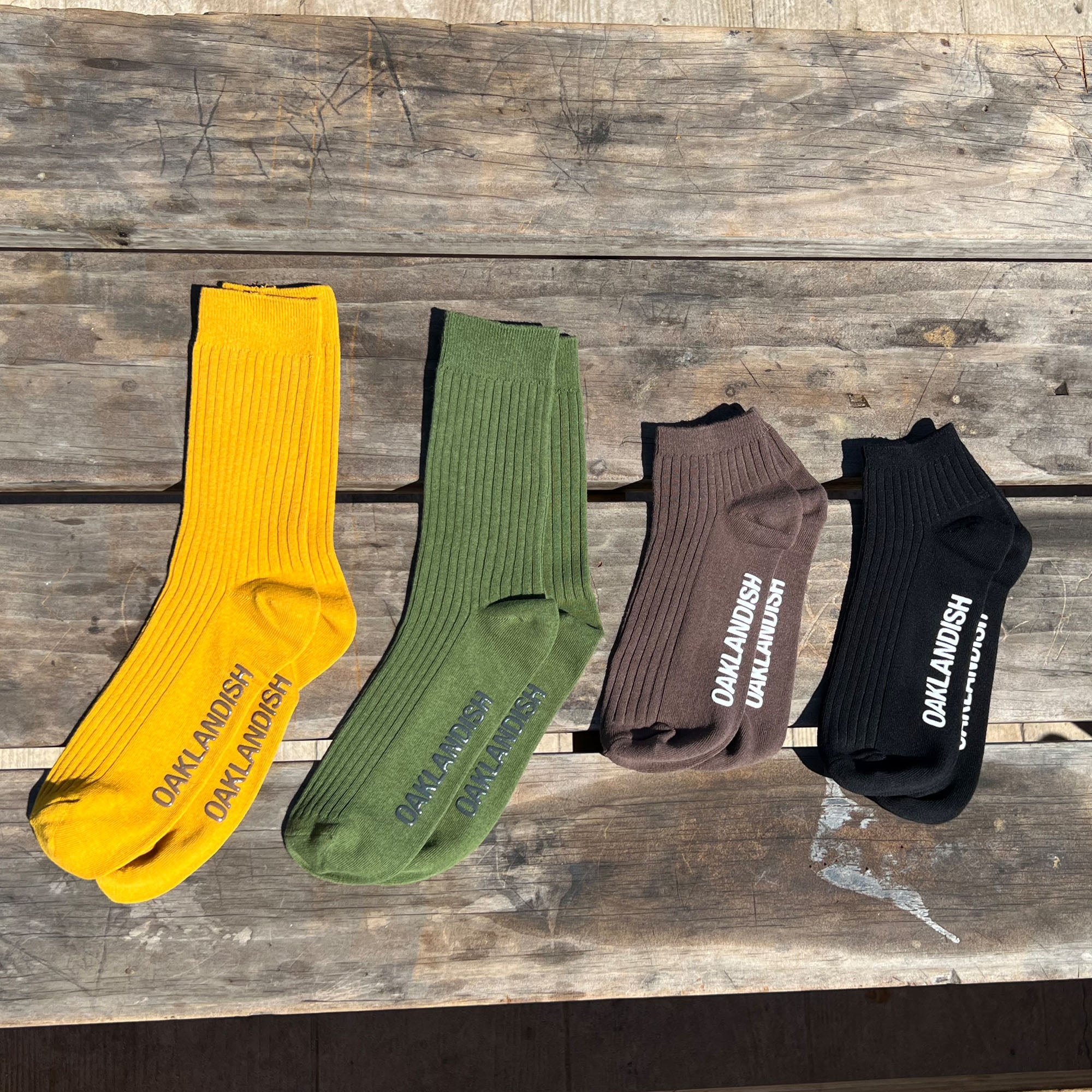 Four pairs of Oaklandish Grip Crew Socks are displayed side by side on a rustic wooden surface. The socks come in different colors: yellow, green, brown, and black. Each pair has "OAKLAND" in white lettering on the sole. Sunlight casts shadows highlighting their texture. One size fits most.